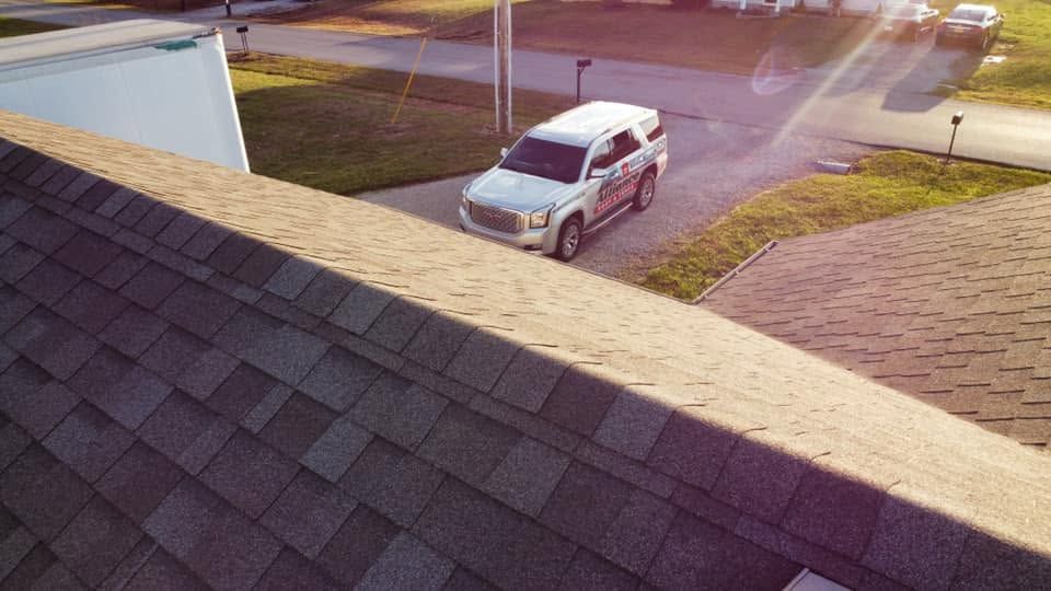 White SUV parked in a driveway beside a house, seen from a shingled roof in warm sunlight