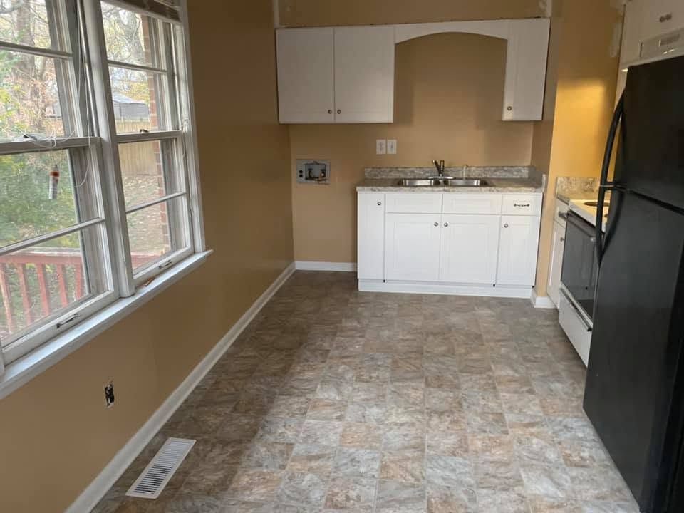 Empty kitchen with white cabinets, tan walls, tile floor, large window, and black refrigerator.