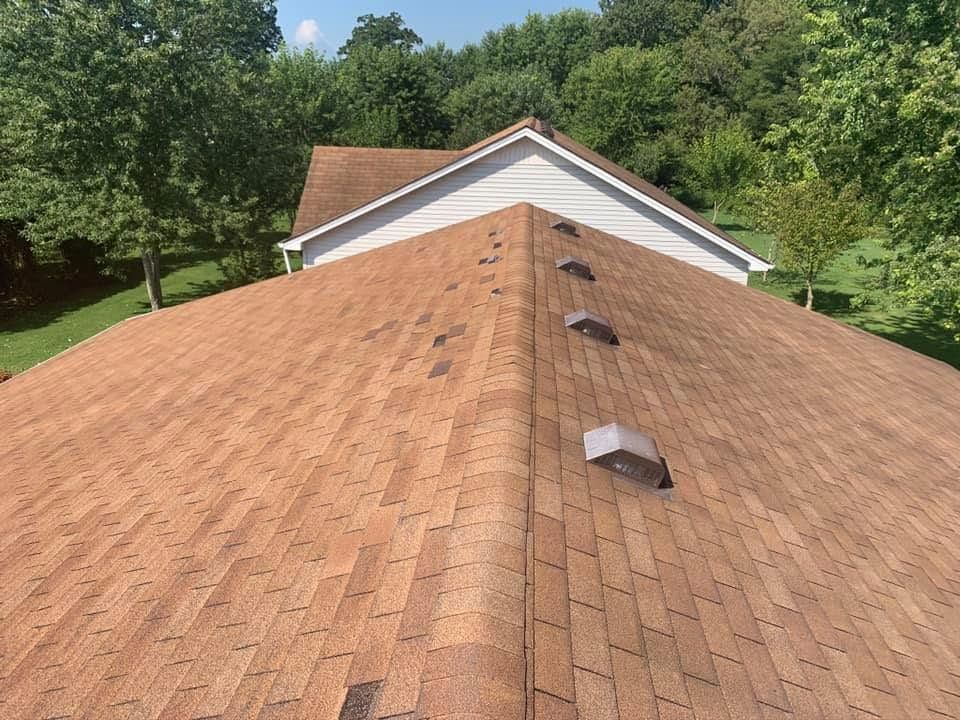 Brown shingled roof with vents, viewed from above toward a white house and green trees