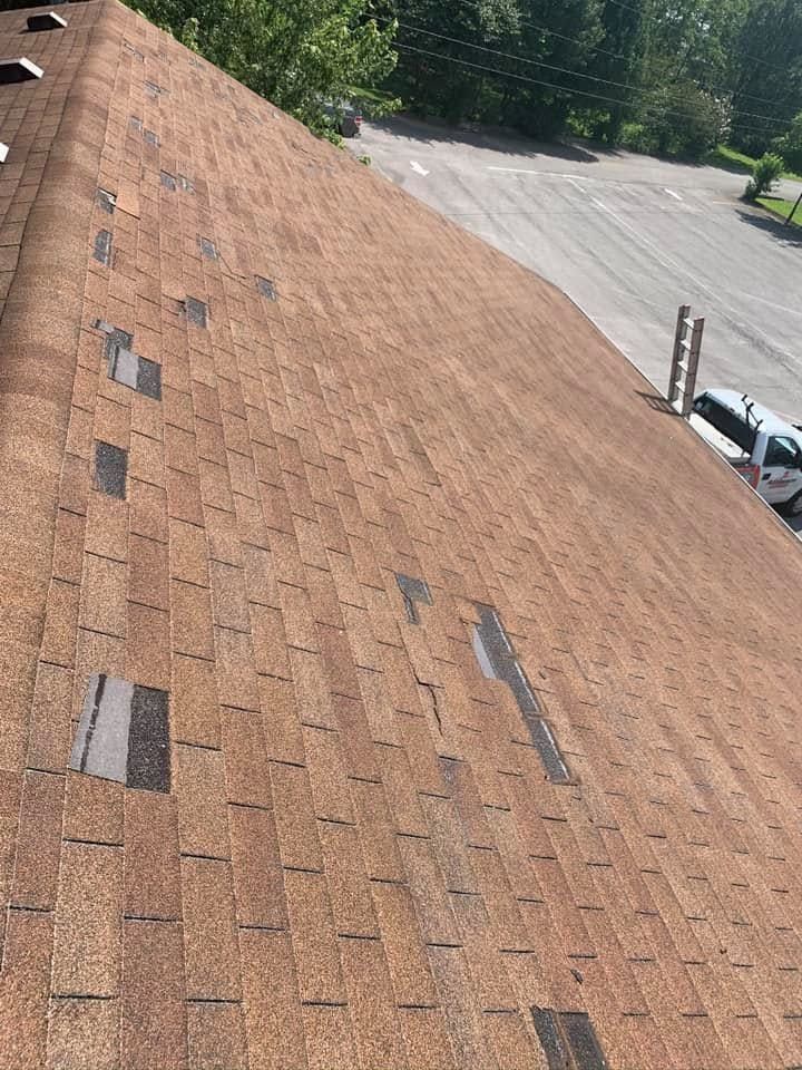 Weathered brown shingle roof with several missing or damaged shingles, viewed beside a parking lot.