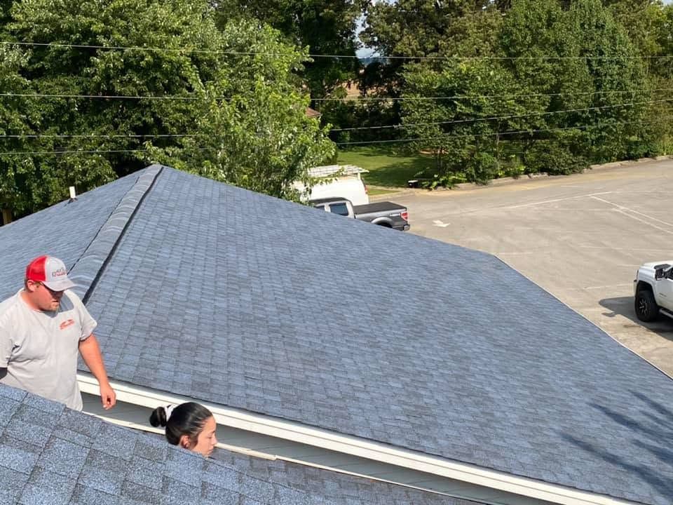 Two workers on a shingled roof beside a parking lot and trees