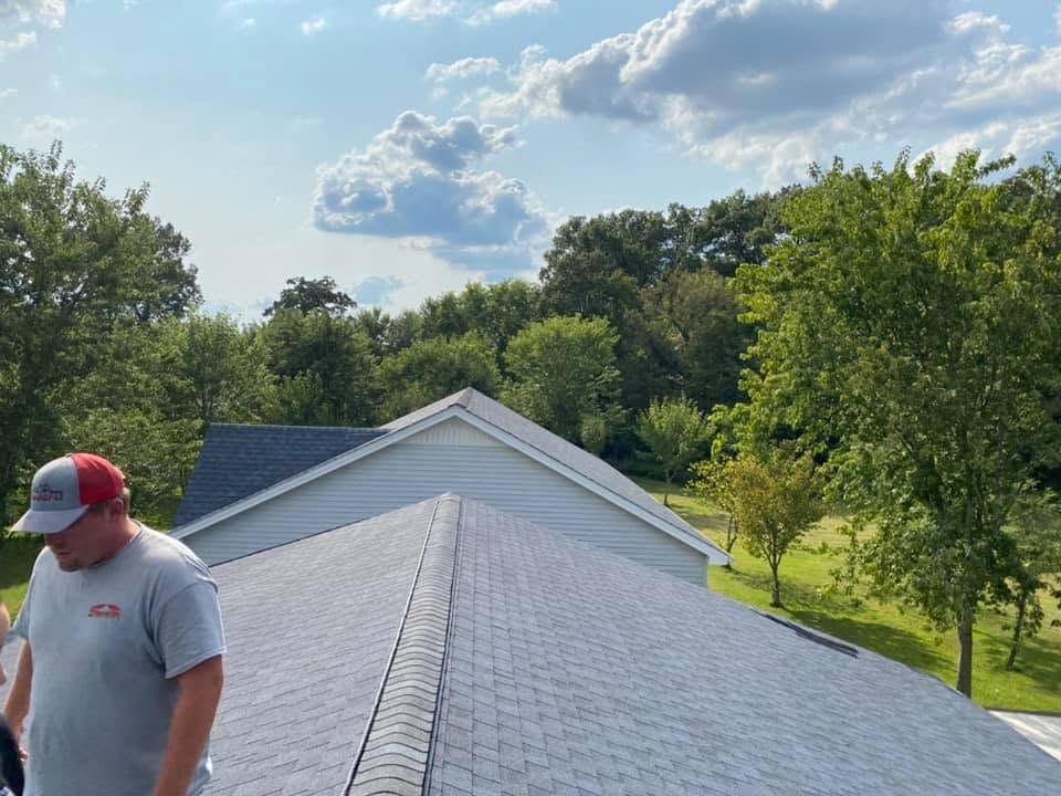 Man on a roof installing gray shingles beside trees under a blue sky