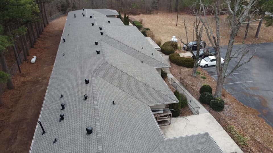 Aerial view of a gray-shingled roof with multiple vents, beside trees, parked cars, and a road.