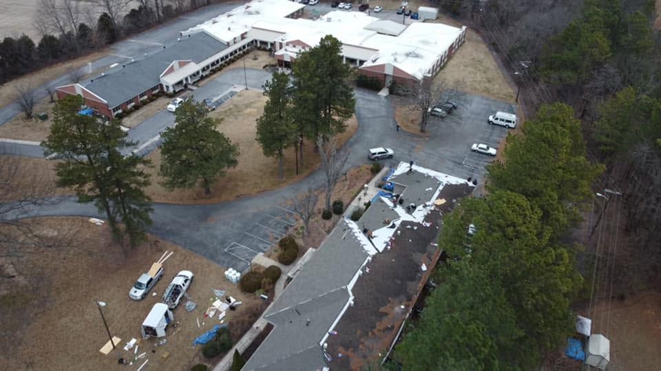 Aerial view of a school campus with white buildings, trees, and parked vehicles on a wet lot.