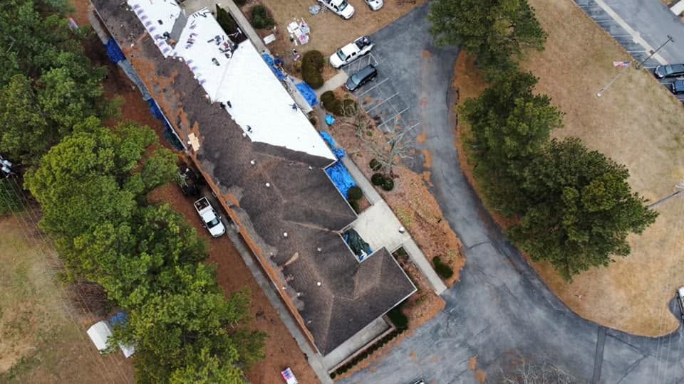 Aerial view of a long building with a white roof beside a gravel lot and trees.