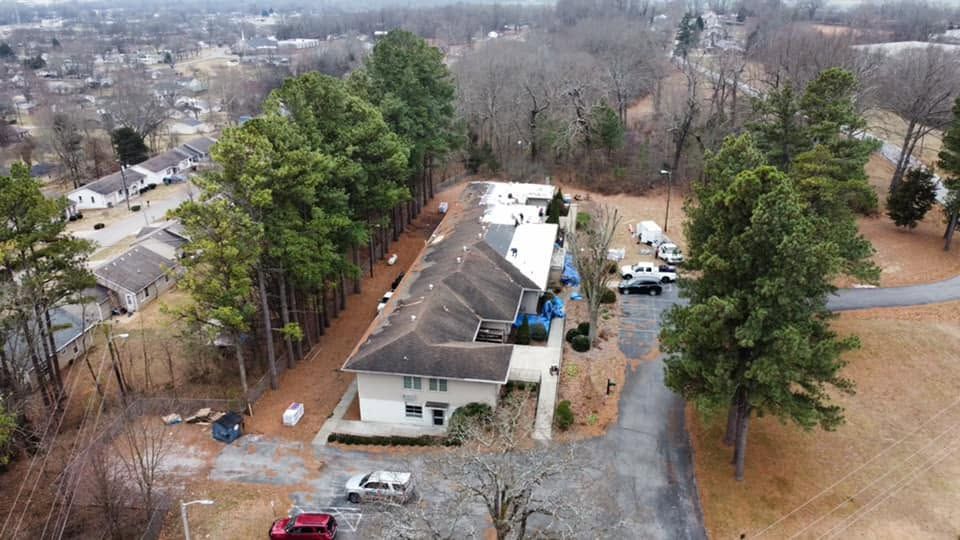Aerial view of a house under repair beside tall trees and a driveway in a wooded neighborhood.