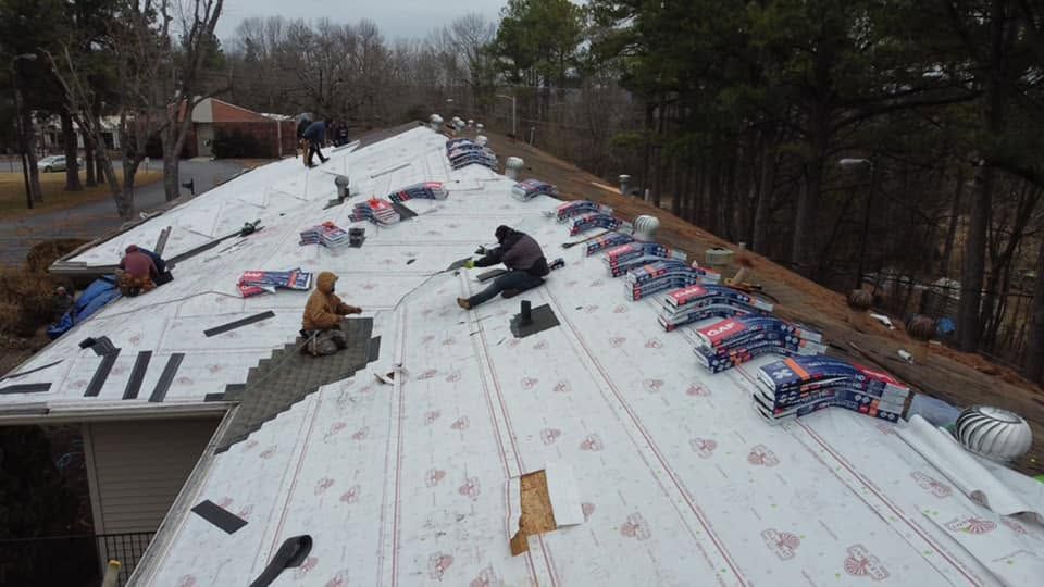 Workers installing white roofing sheets on a long roof, with tools and materials scattered along it.