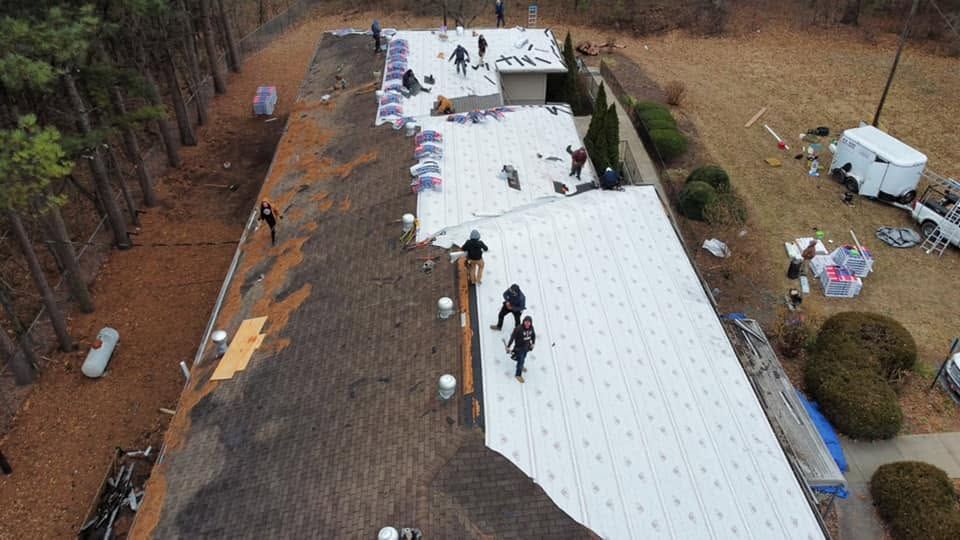 Workers installing white roofing panels on a long roof in a wooded area, with tools and materials nearby