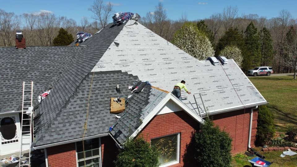 Workers reroofing a brick house, with dark shingles on one side and bare roof deck on the other.