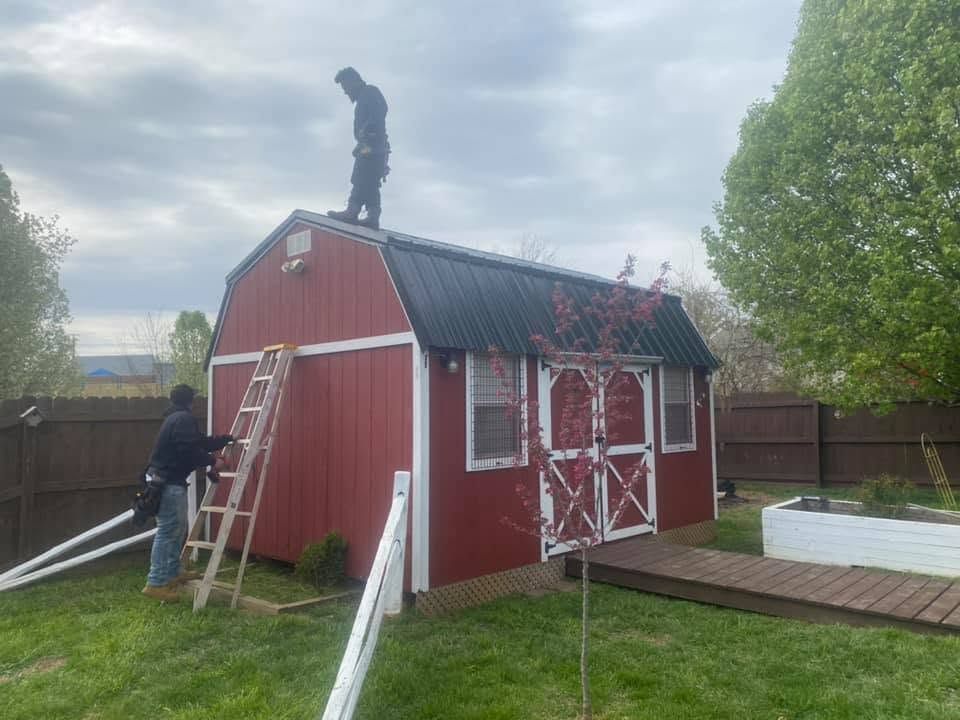 Person on roof of red barn using a ladder; another stands by it in a yard.