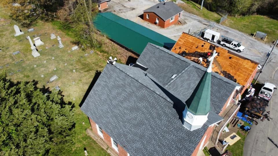 Aerial view of a house with gray shingles, green-roofed building, and yard beside a road