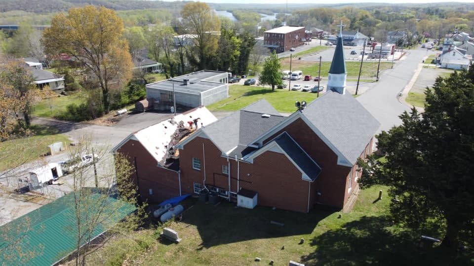 Aerial view of a red-brick church with a gray roof beside a quiet street and trees.