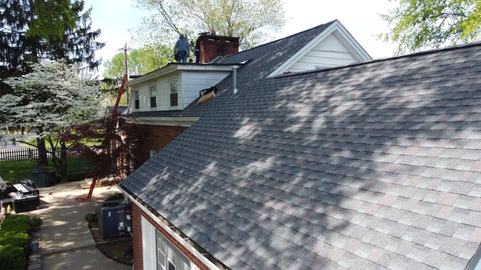 Suburban house roof with gray shingles, dormer windows, and trees in the yard