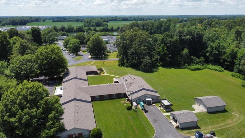 Aerial view of a rural building complex with green lawns, trees, and open fields under a cloudy sky