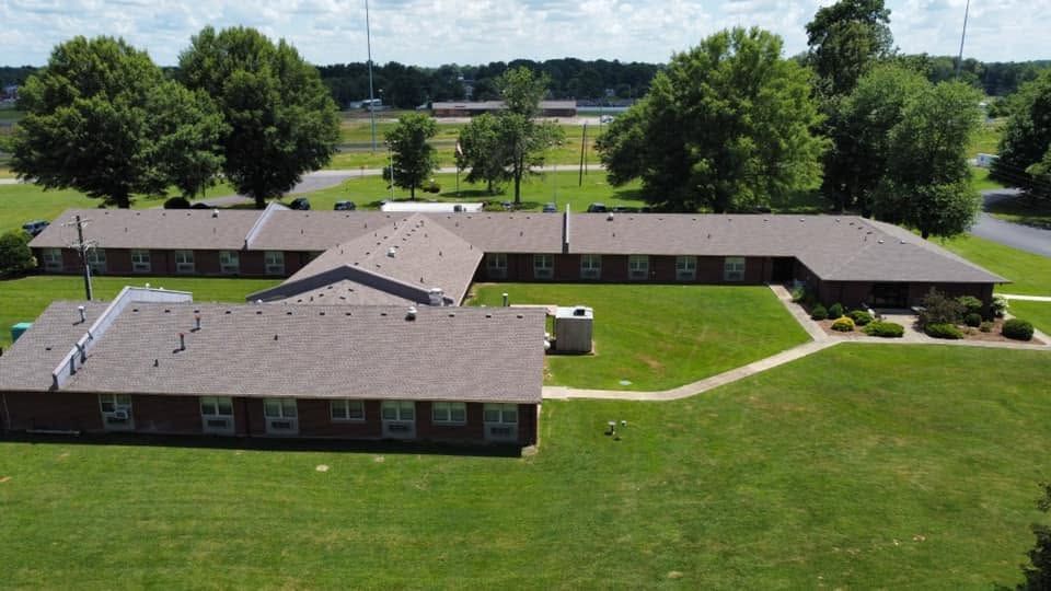 Aerial view of a single-story brick building with a gray roof, surrounded by green lawn and trees.