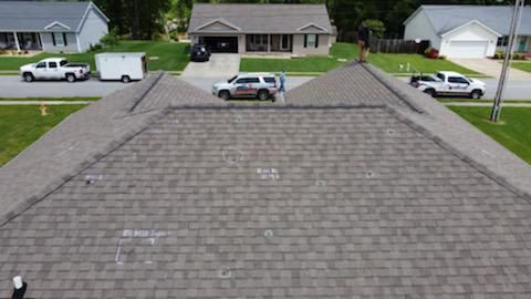 Aerial view of a gray shingled roof with houses and parked vehicles visible across the street.