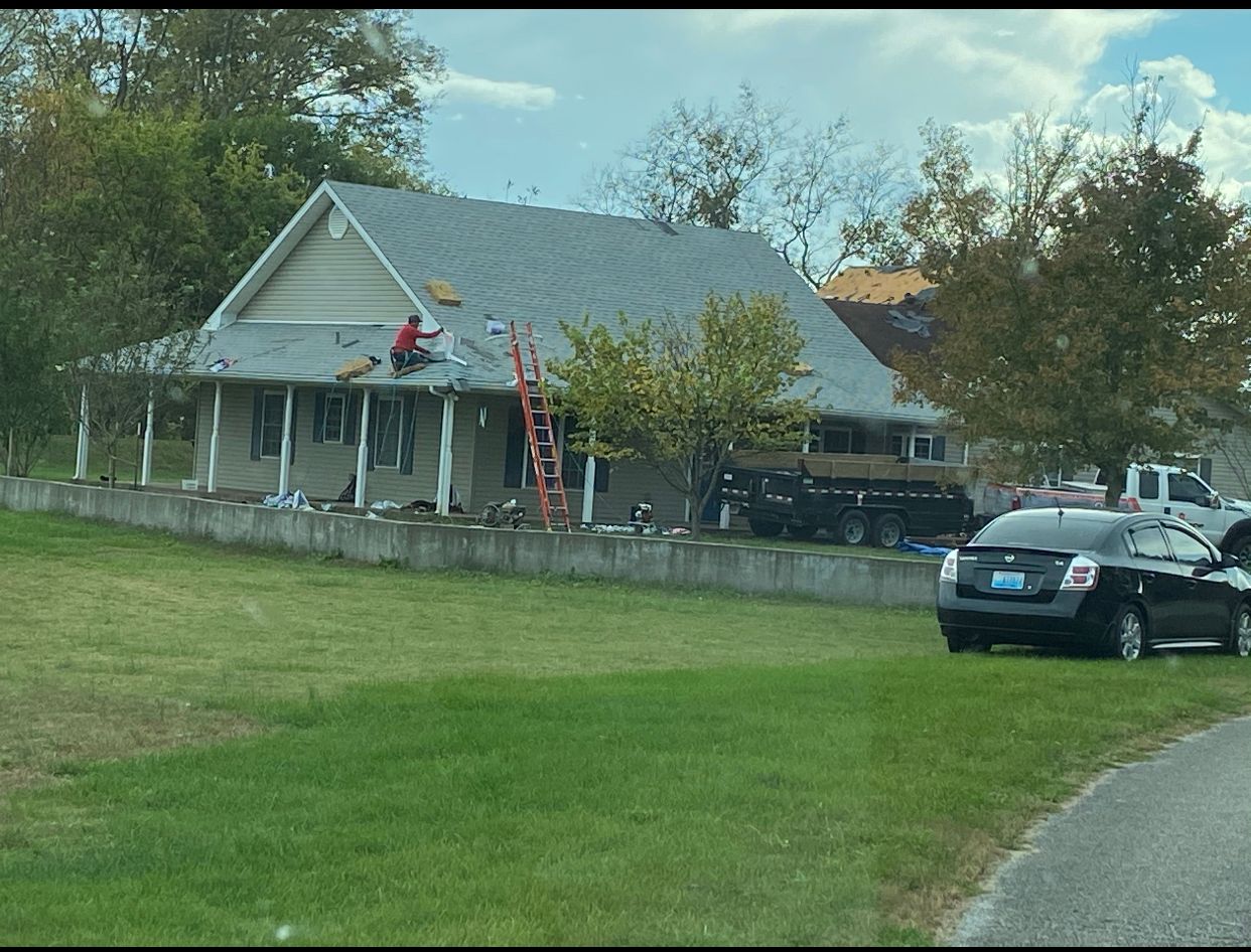 House with a gray roof, ladder, and workers in a yard beside parked cars.
