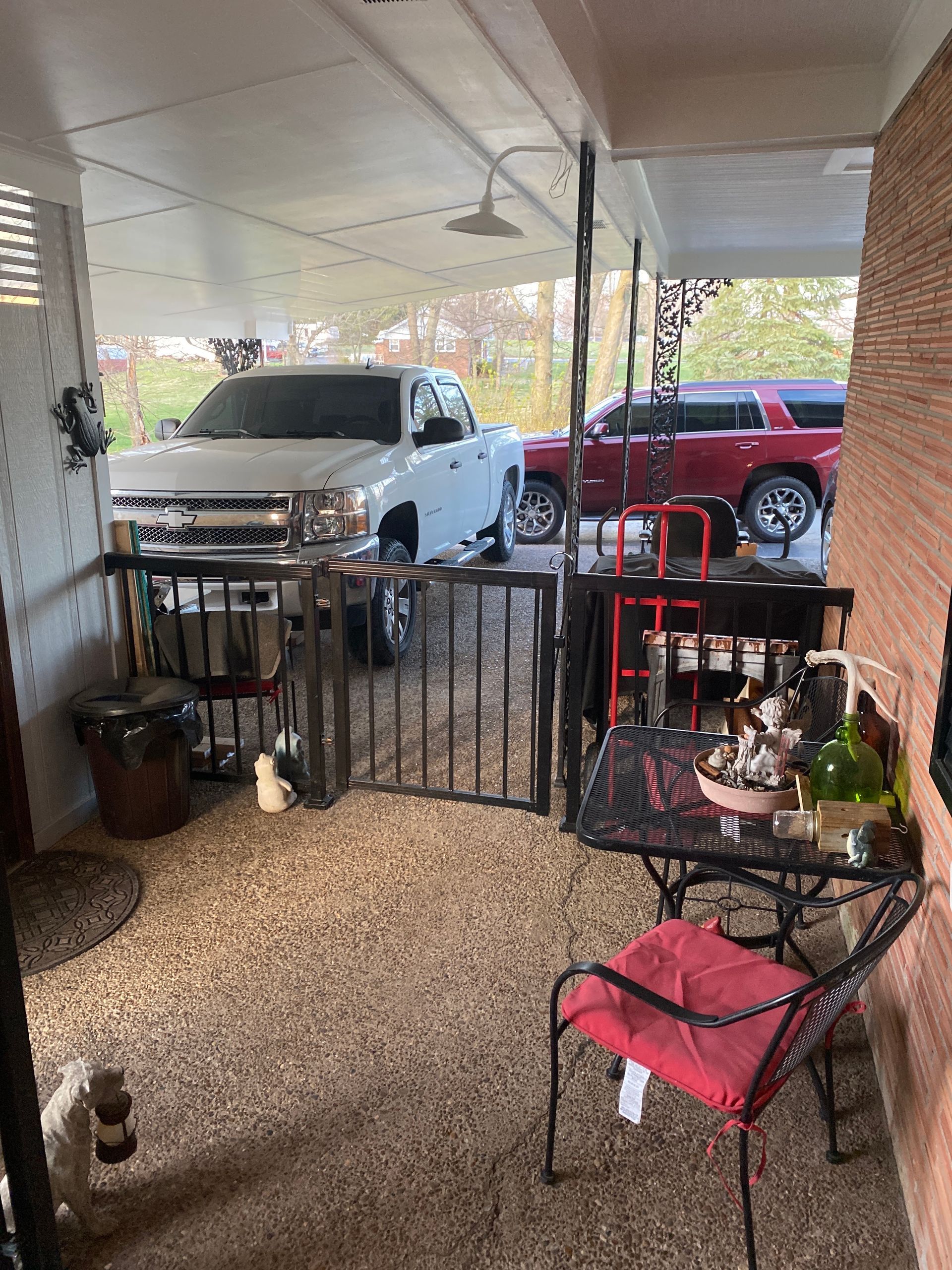 Front porch with metal gate and chairs, overlooking parked white and red trucks under a covered carport.