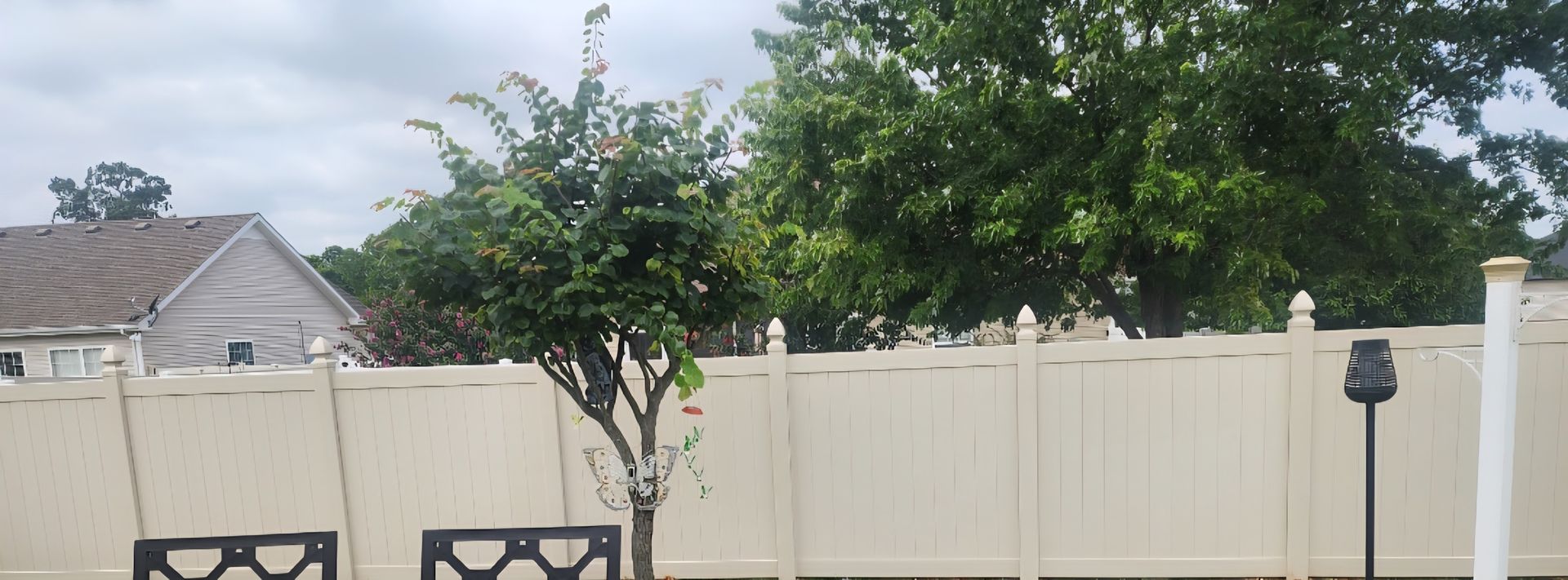 Backyard with beige vinyl fence, small trees, patio chairs, and a house roof in the background