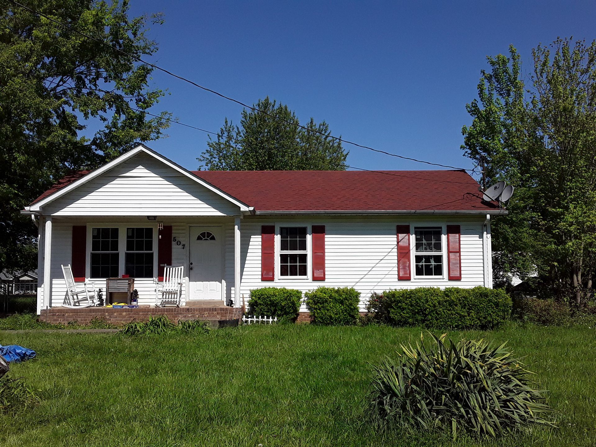 Single-story white house with red roof and shutters, front porch, and green lawn under a clear blue sky