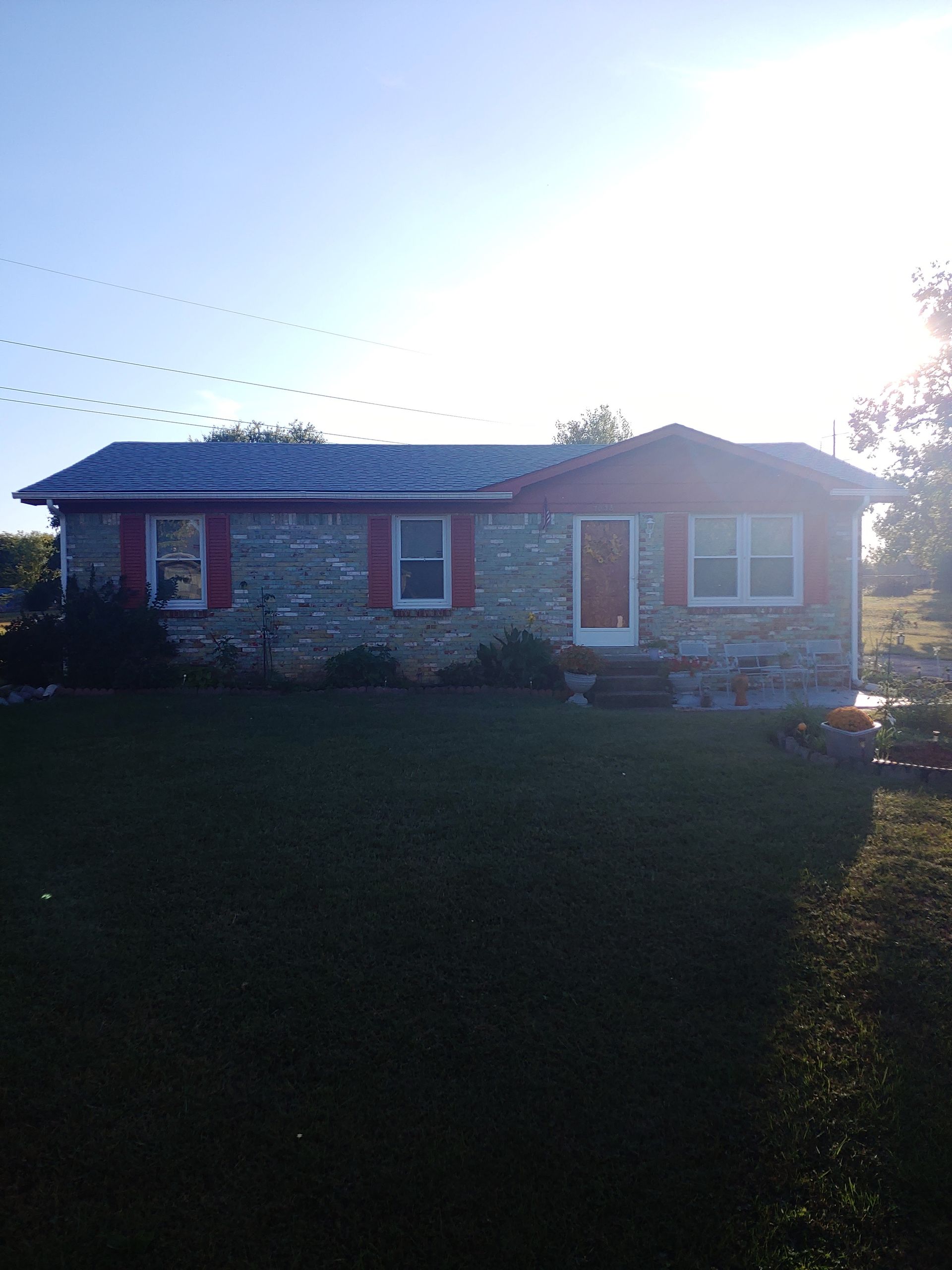 Brick house with red shutters in bright sunlight, viewed from a dark front yard.