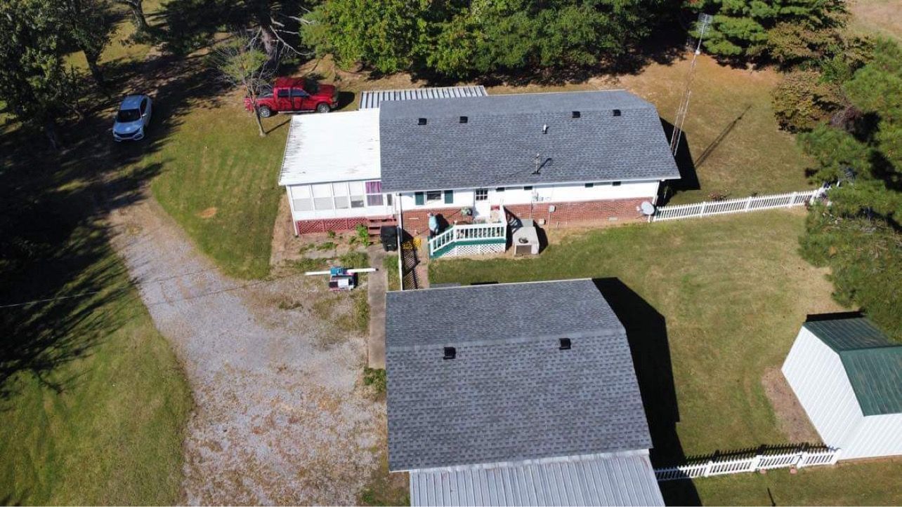 Aerial view of two white houses with gray roofs, gravel driveways, and a red car near trees.