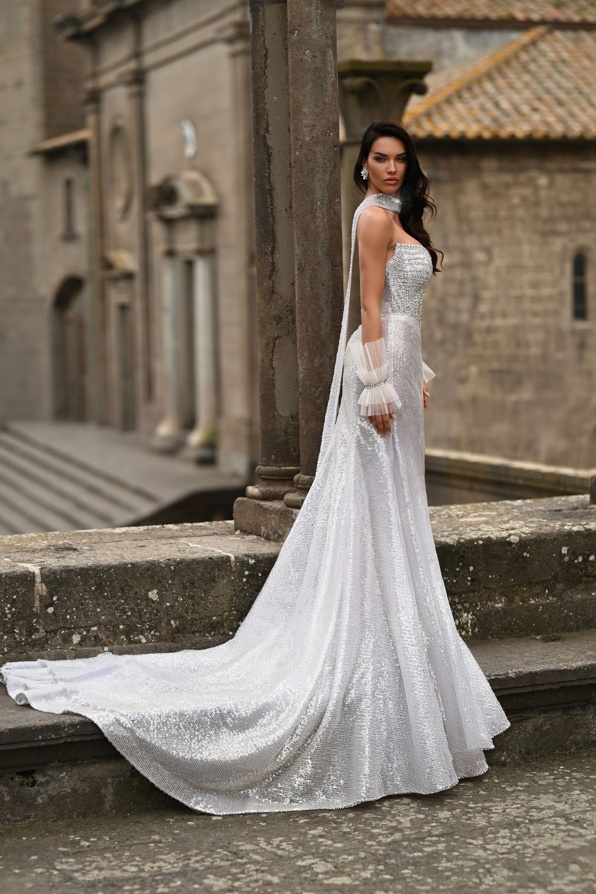 Woman in a white lace gown posing outdoors by stone columns, with a long trailing train.