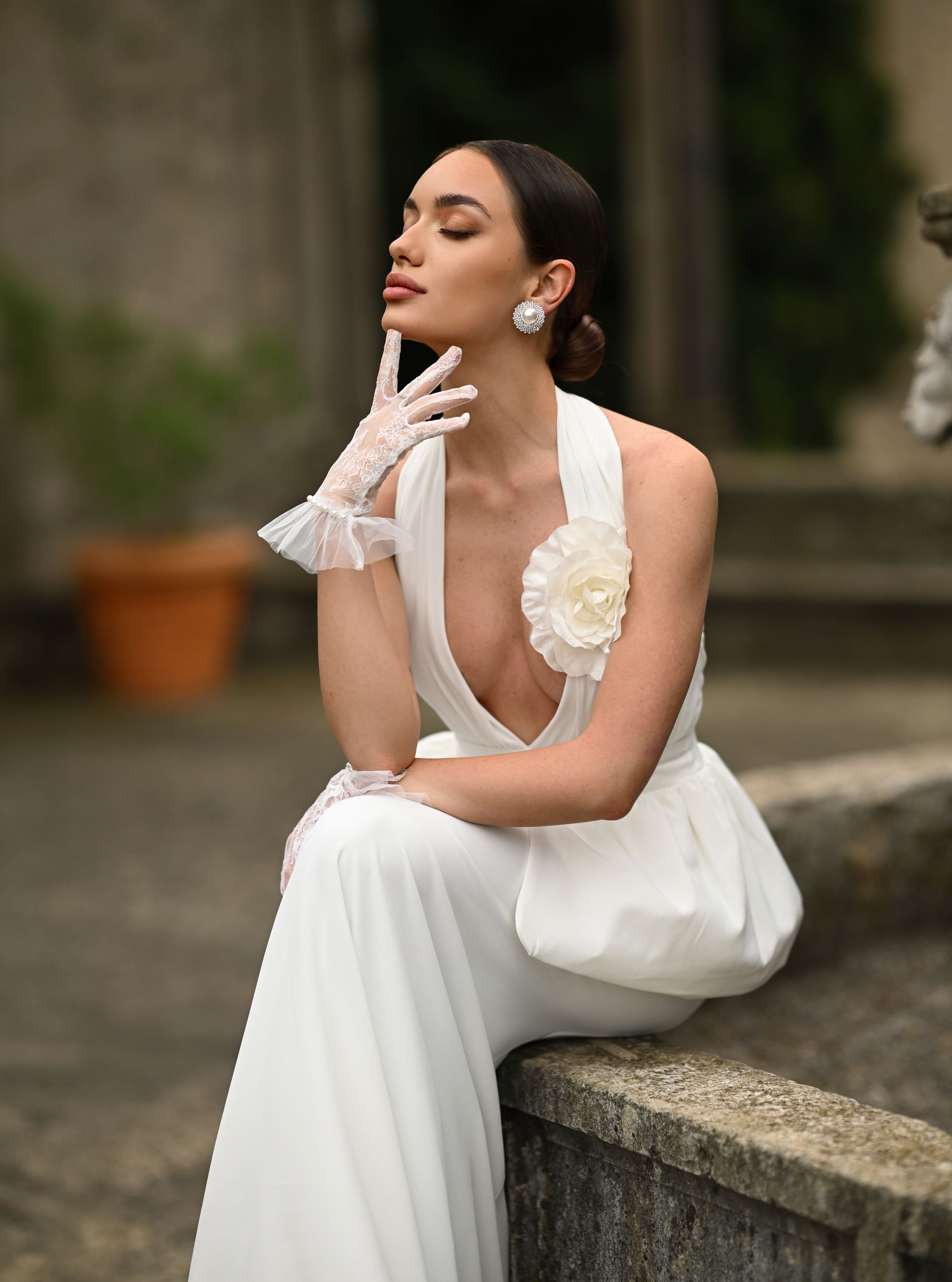 Woman in a white gown and gloves sitting on a stone ledge in a garden, posing thoughtfully.