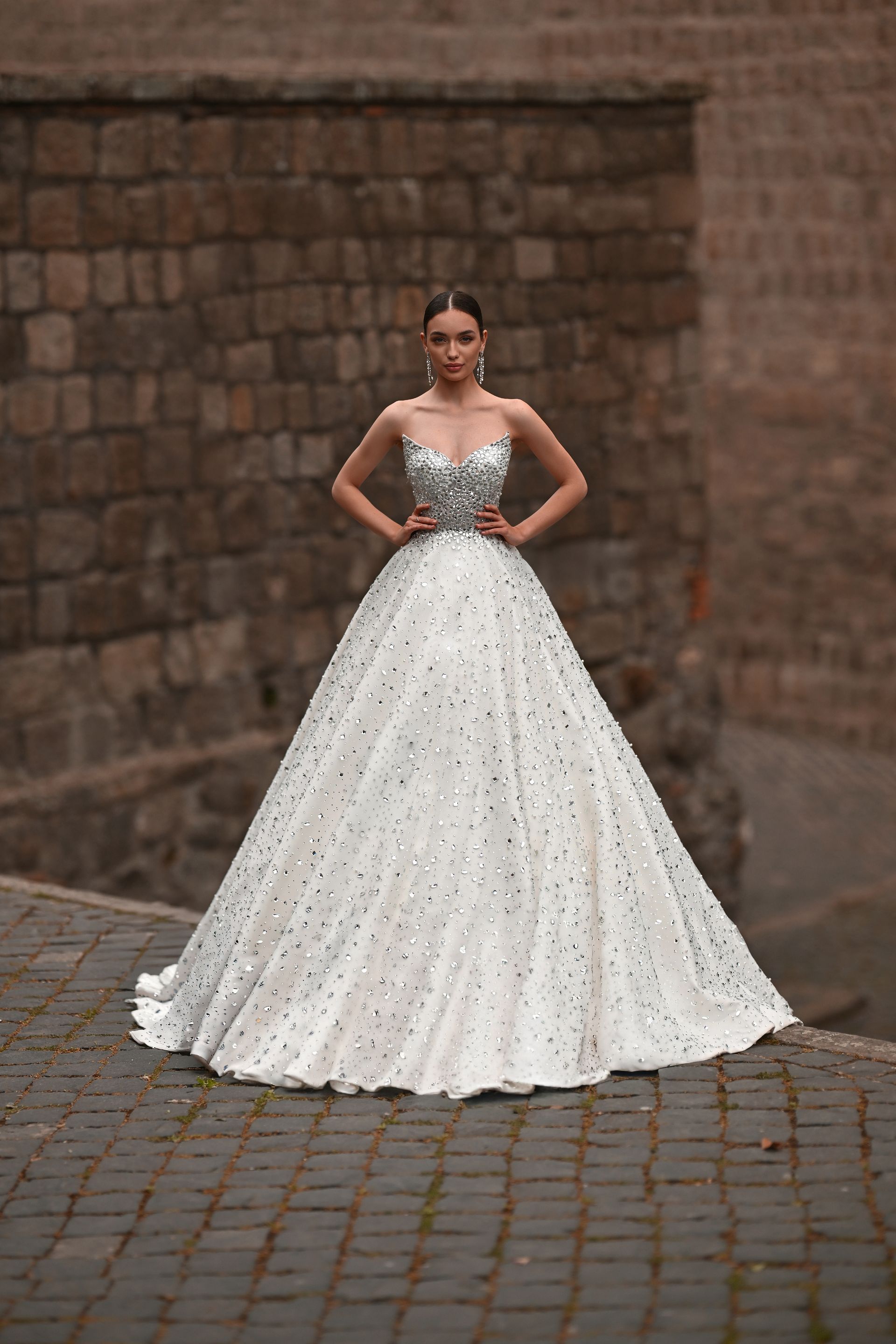 Woman in a white ball gown with a silver bodice, standing on cobblestones against a brick wall.