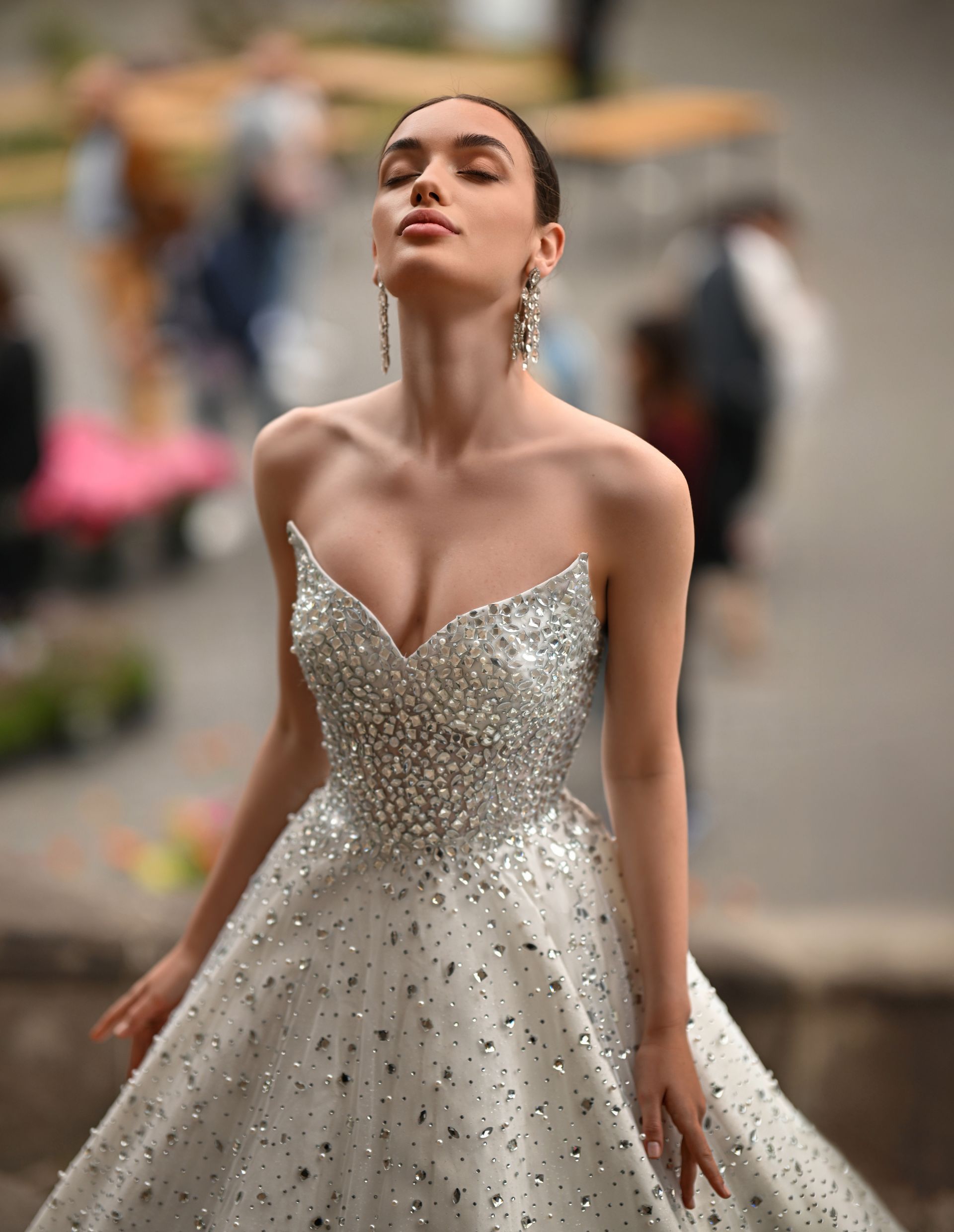 Woman in a sparkling white gown posing outdoors with eyes closed, wearing dangling earrings.