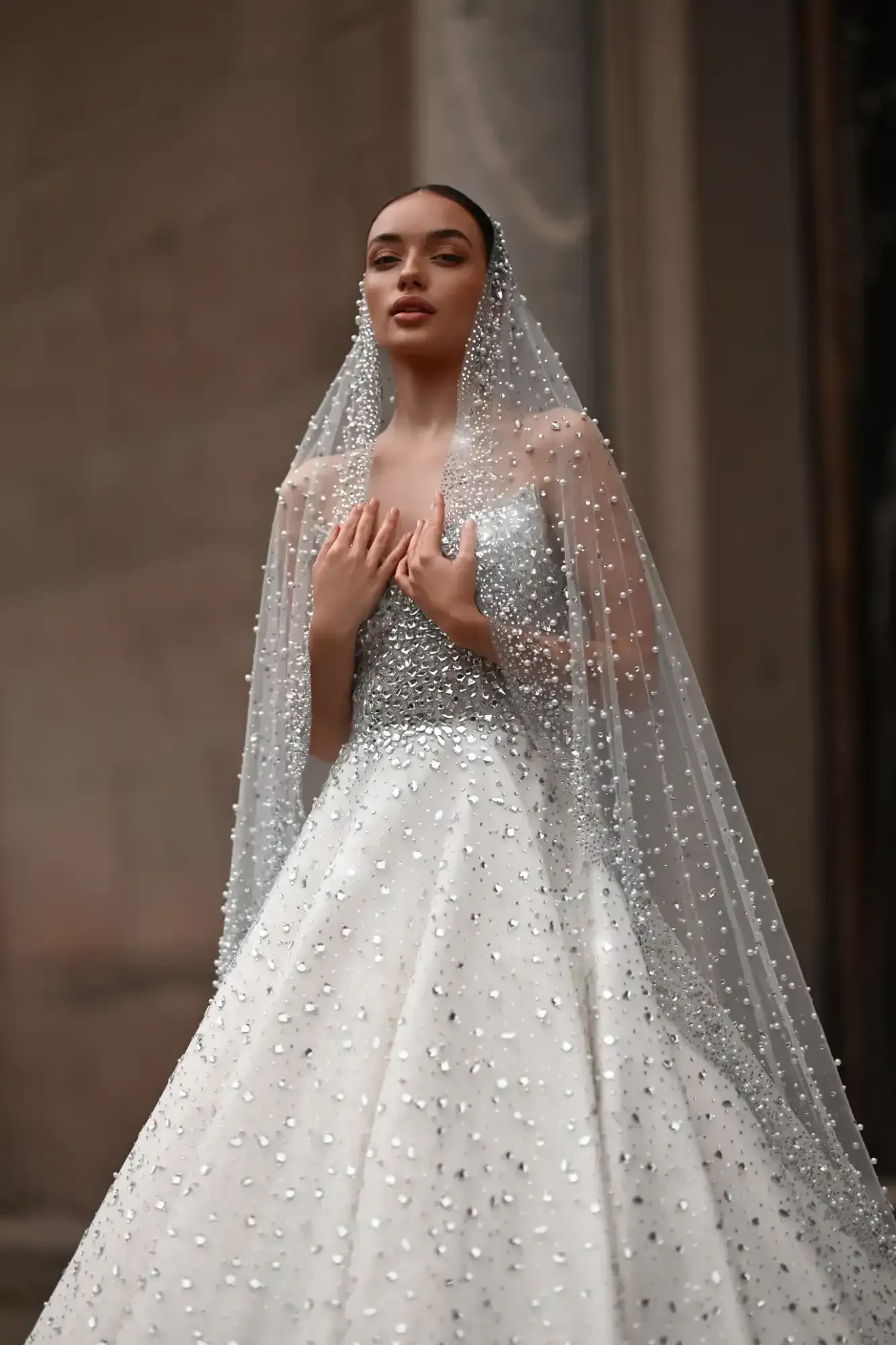 Woman in a sparkling white gown and veil standing indoors with hands clasped at her chest.