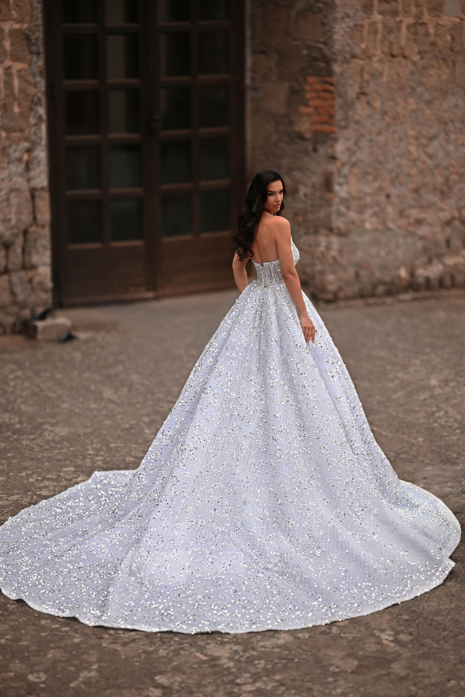Woman in a sparkling white ball gown posing outdoors against a rustic stone wall.