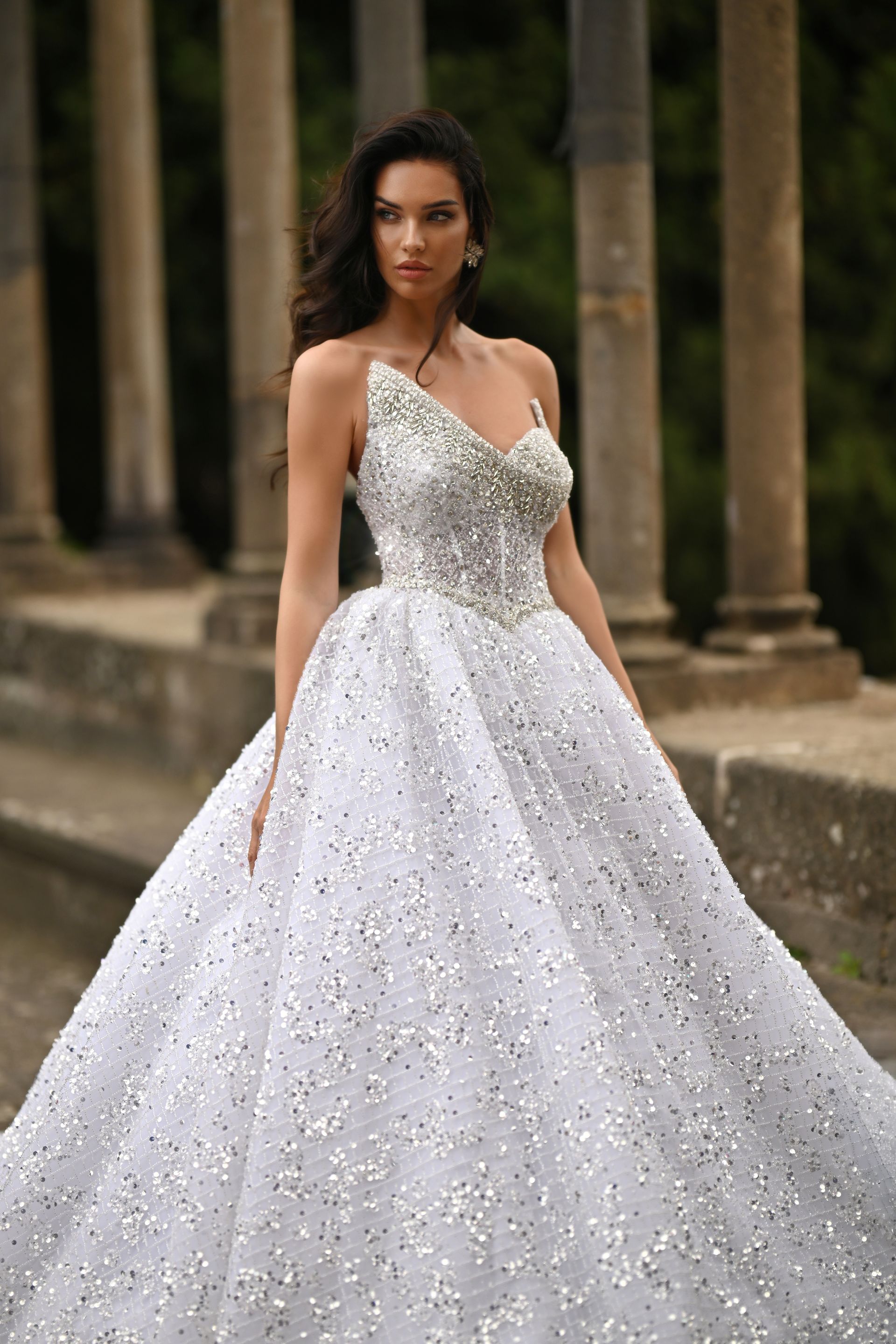 Woman in a sparkling white ball gown standing outdoors among stone columns.