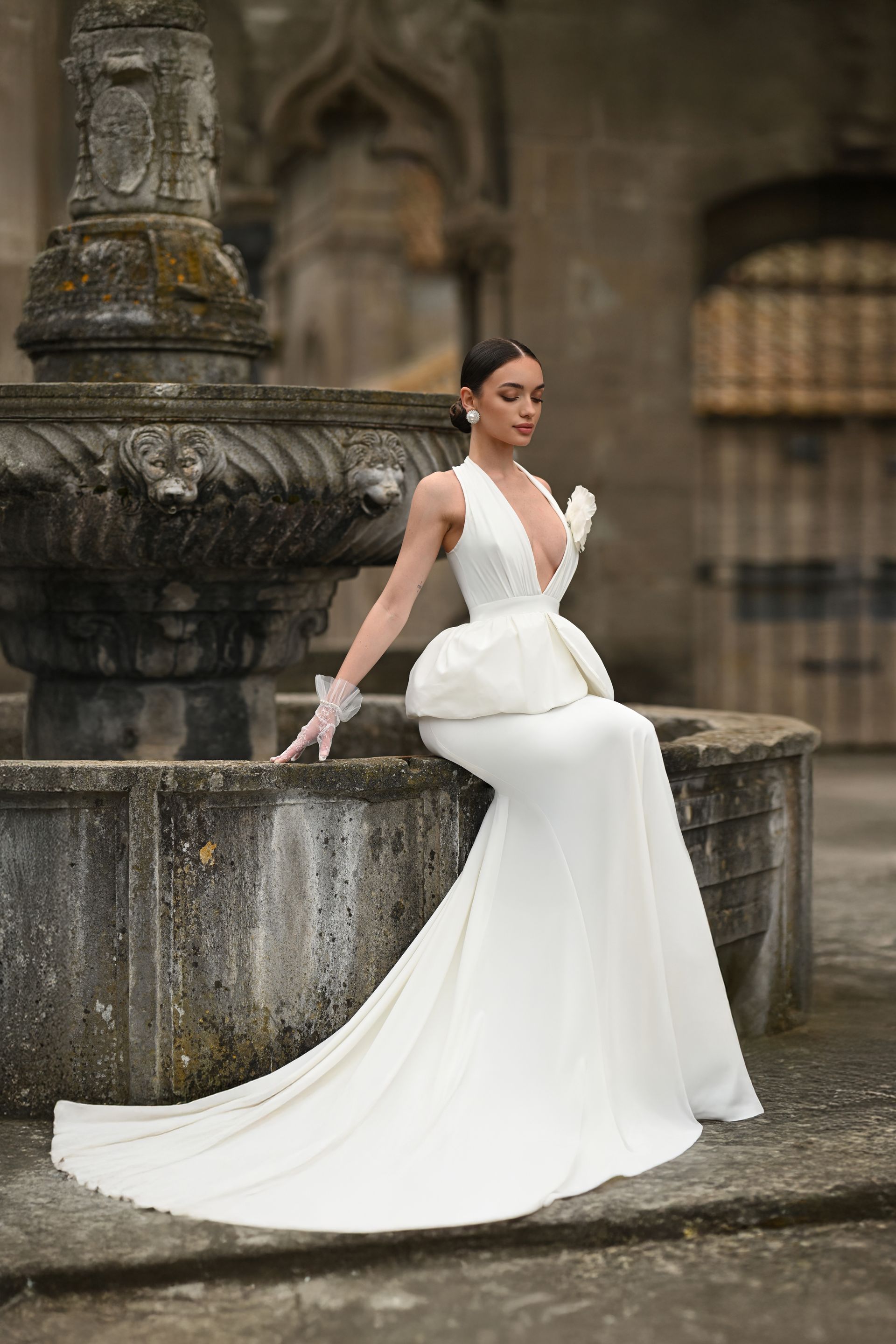 Woman in a white gown sitting on a stone fountain in a historic courtyard.