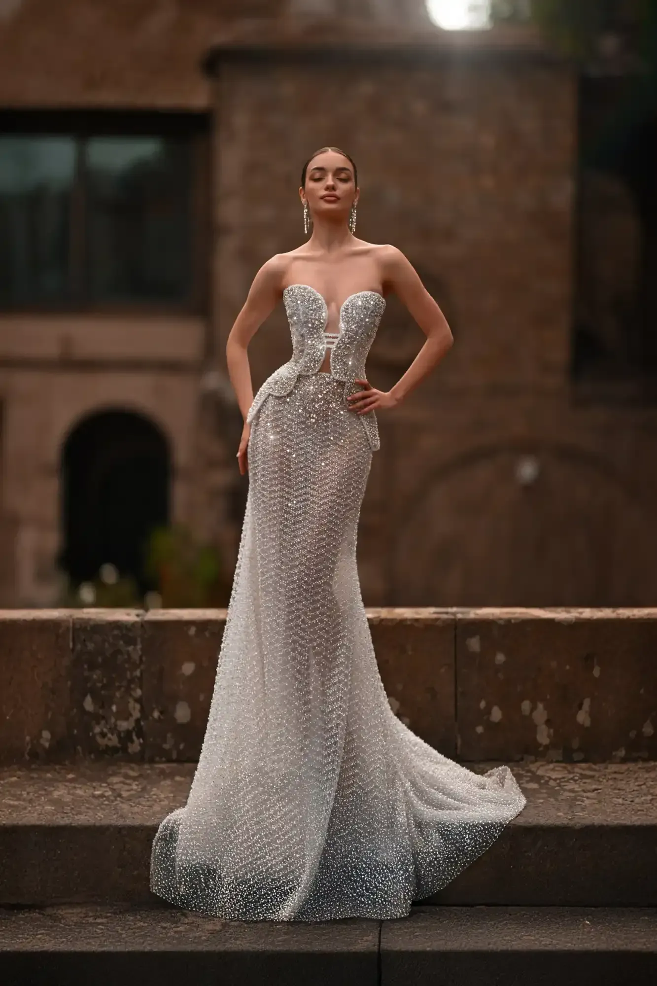 Woman in a sparkling silver gown posing outdoors on stone steps before a brick building.