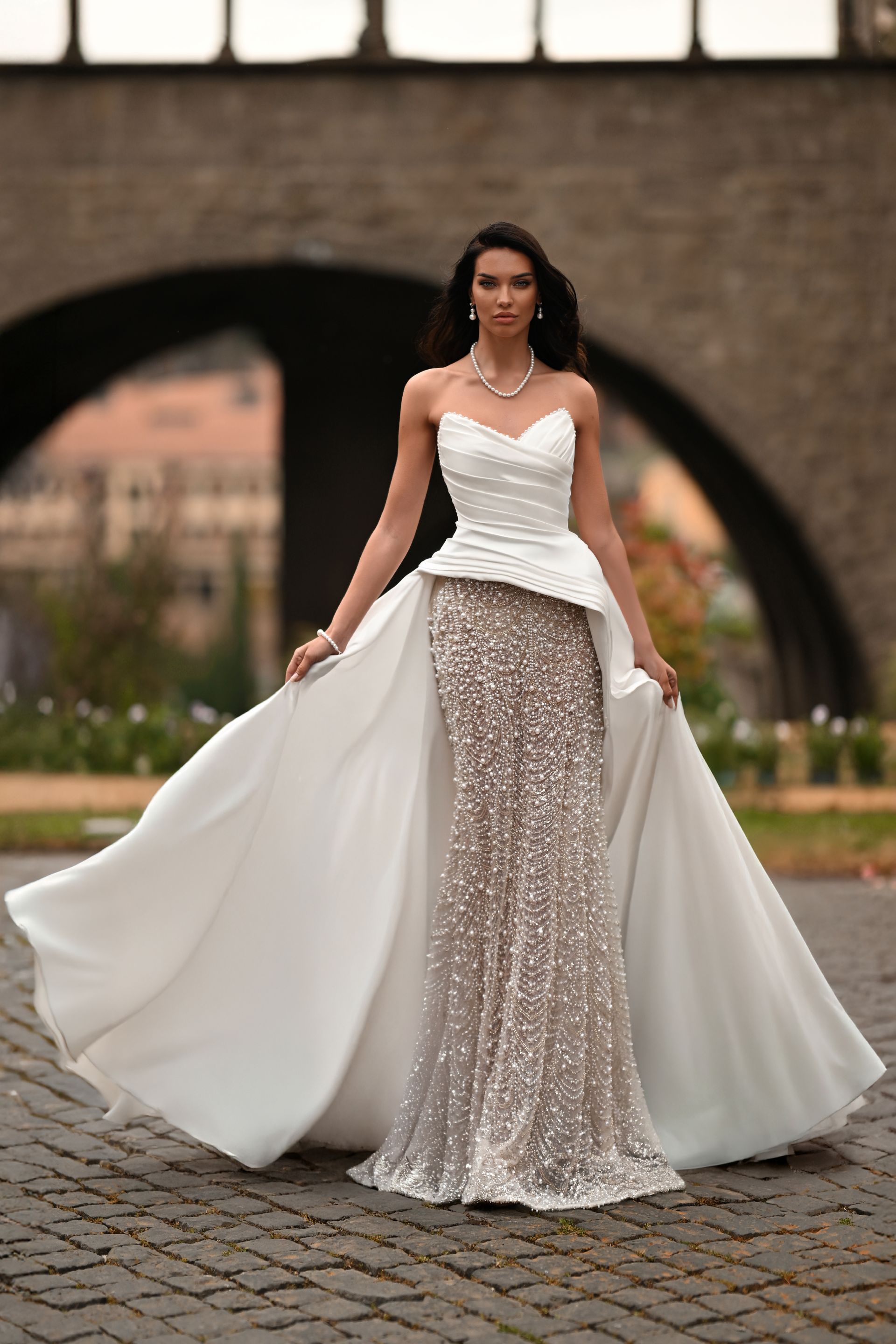 Woman in a white and silver gown standing outdoors with flowing train and arched stone backdrop