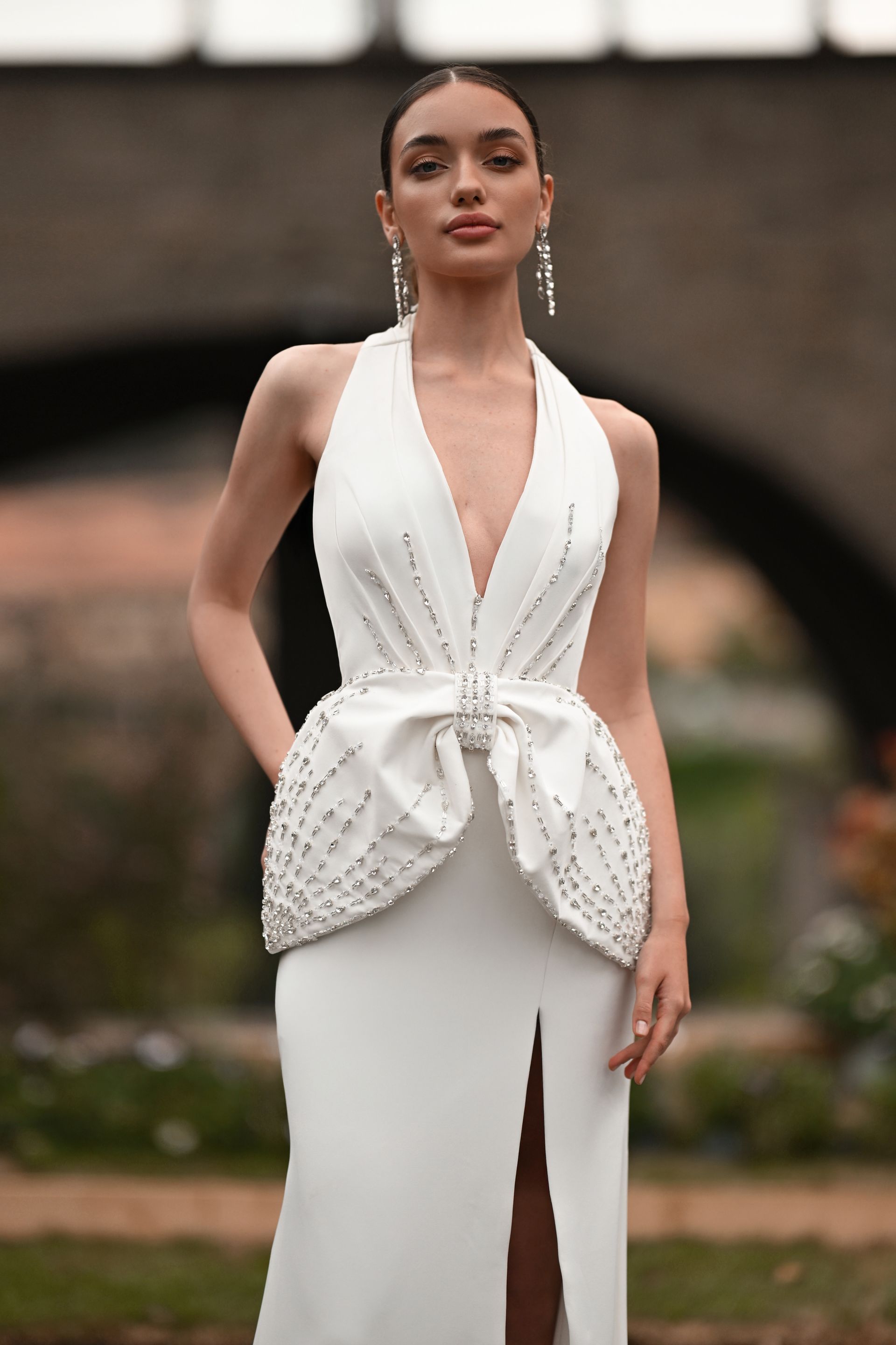 Woman in a white halter gown with a front slit, standing outdoors in a garden setting.