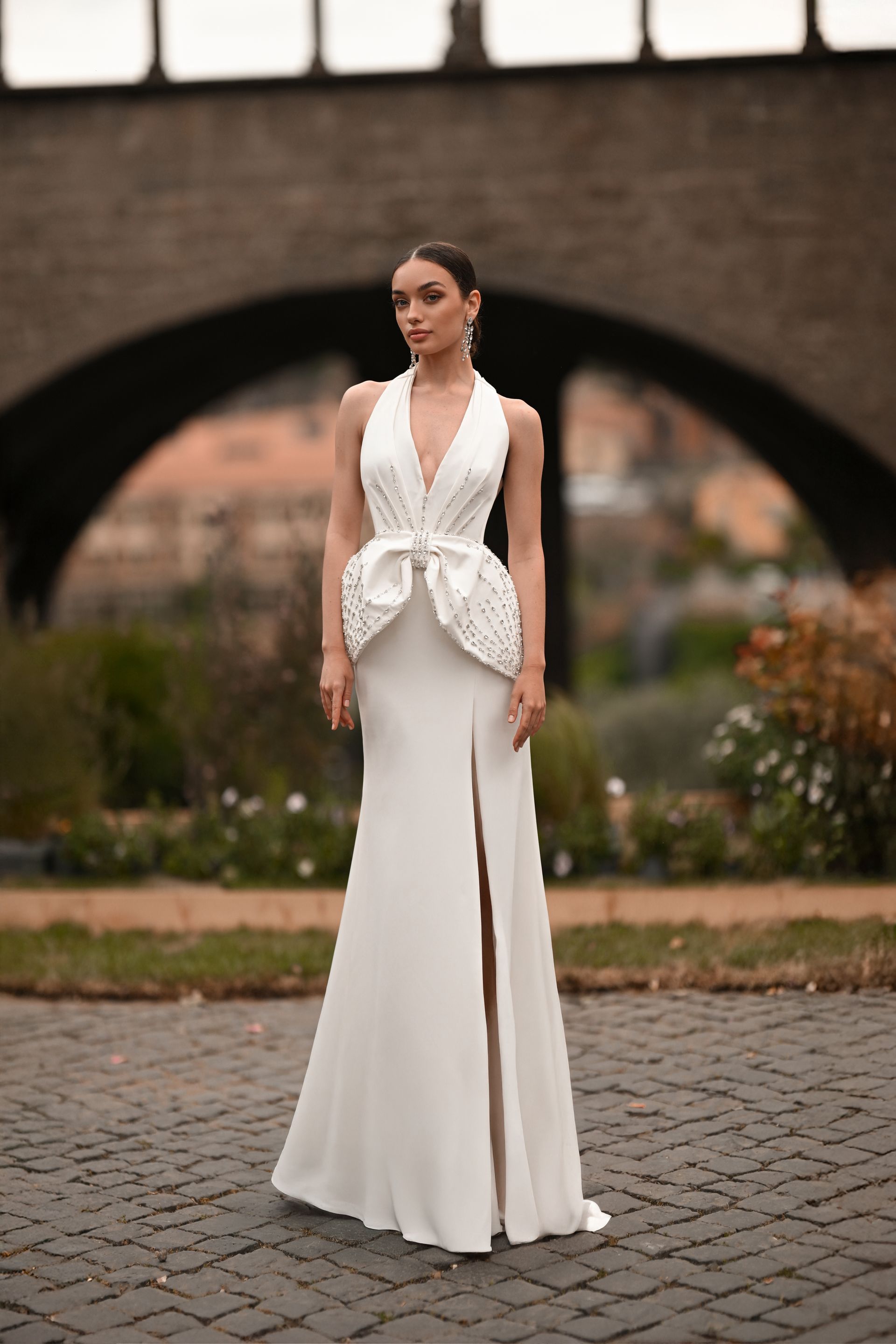Woman in a white halter gown standing on a cobblestone path in a garden, with an arched brick backdrop
