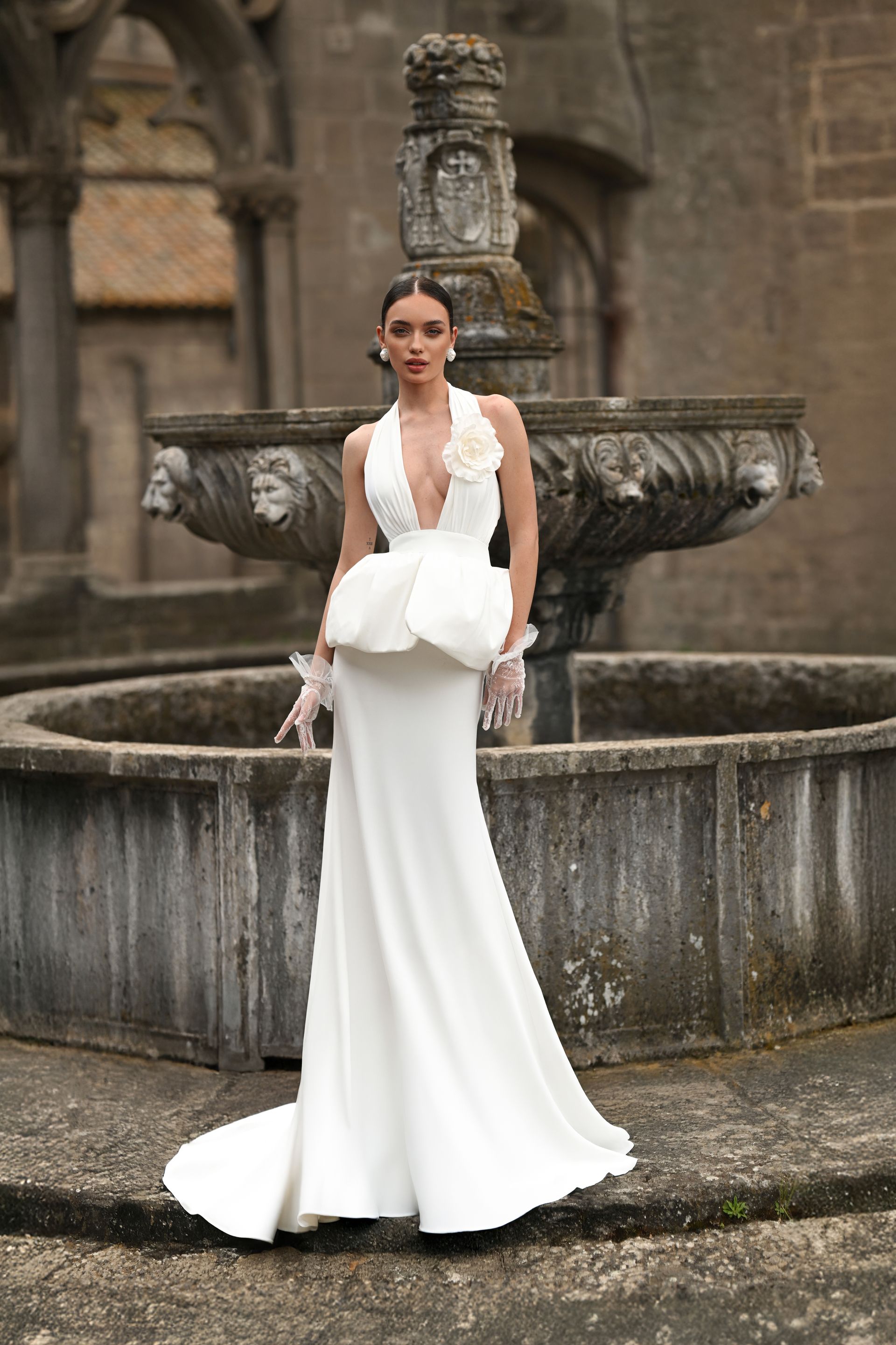 Woman in a white gown standing by an ornate stone fountain in a courtyard