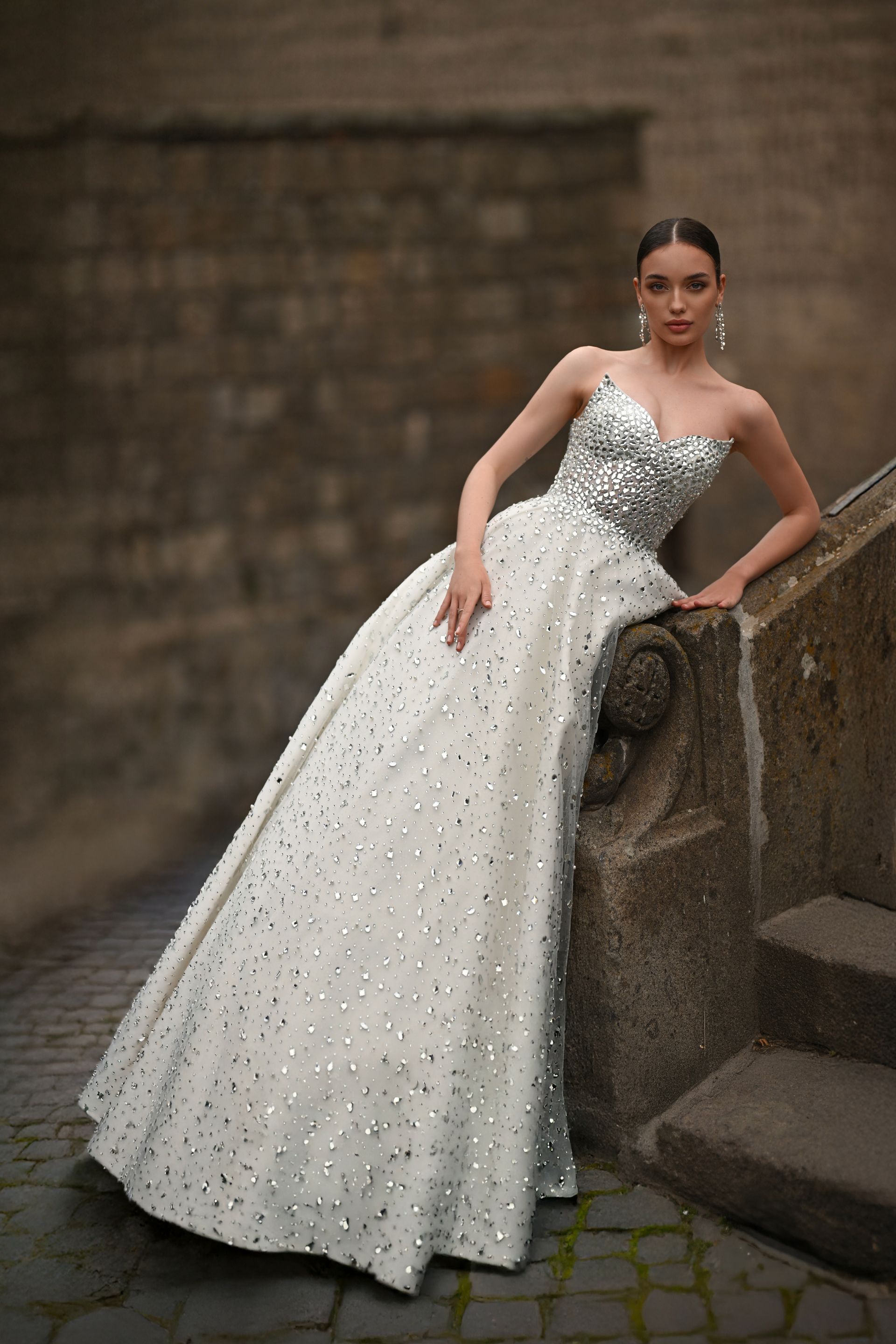 Woman in a white beaded gown leaning on stone steps in a dim courtyard.
