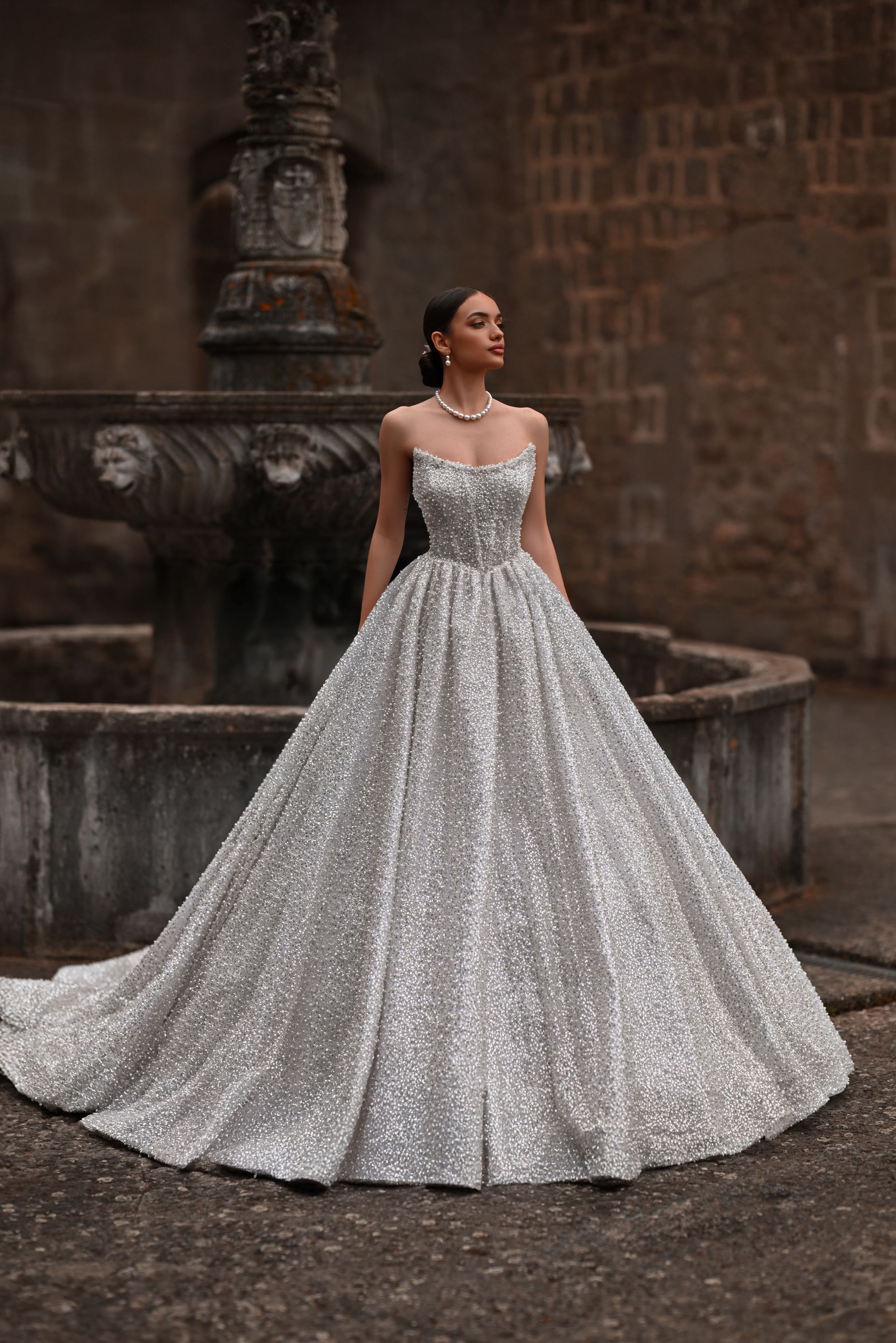 Woman in a silver ball gown standing by a stone fountain in a courtyard