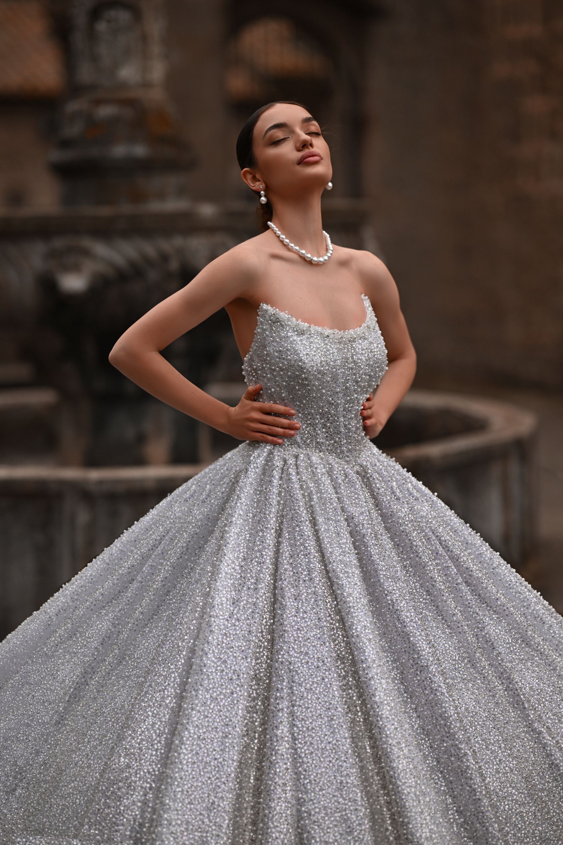 Woman in a sparkling silver ballgown posing outdoors beside a stone fountain