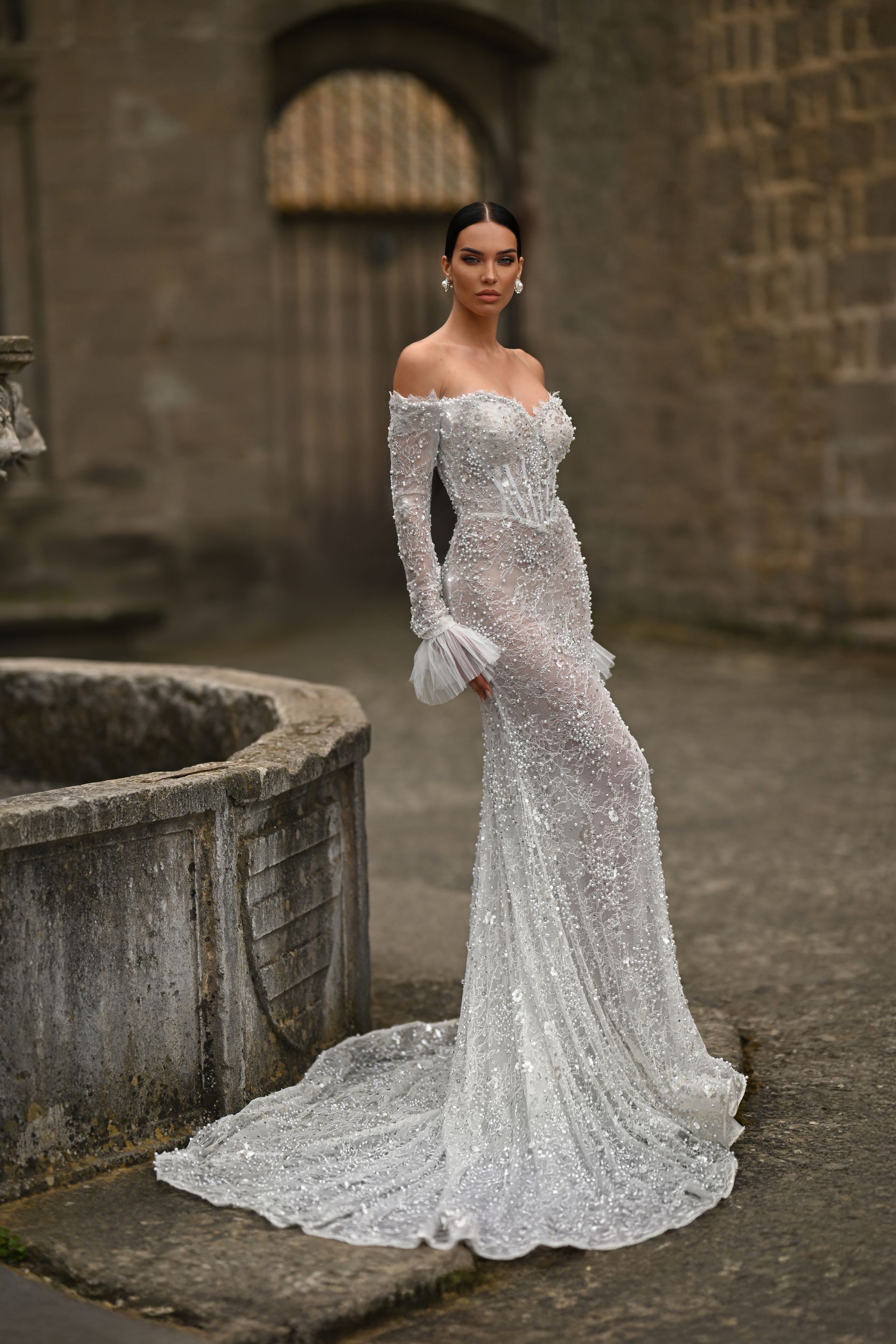 Woman in a sparkling white off-shoulder gown standing in a stone courtyard.