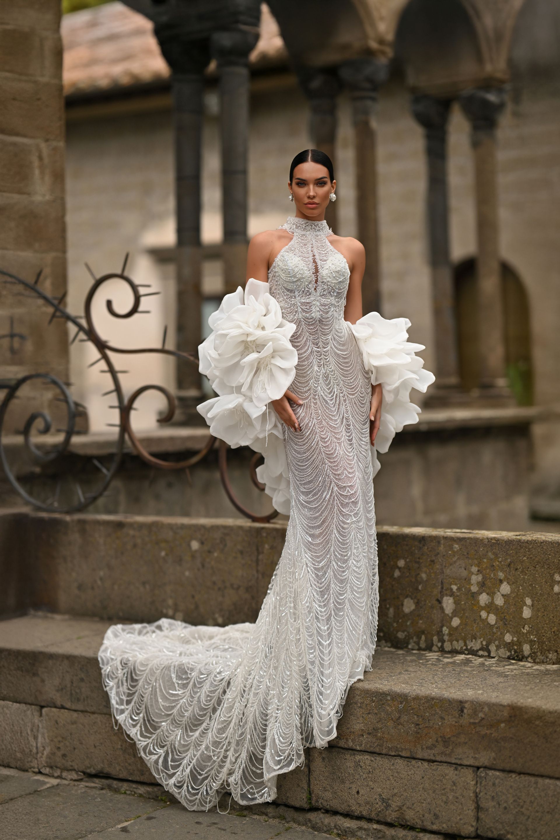 Woman in a silver floral gown with large white sleeves, standing on stone steps in a courtyard.