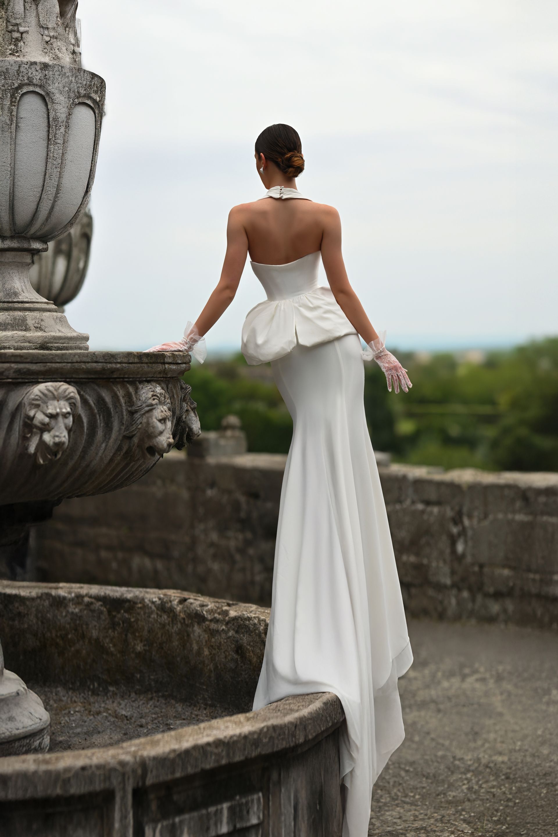 Woman in a white gown standing on stone steps by a terrace, facing away toward a cloudy landscape.