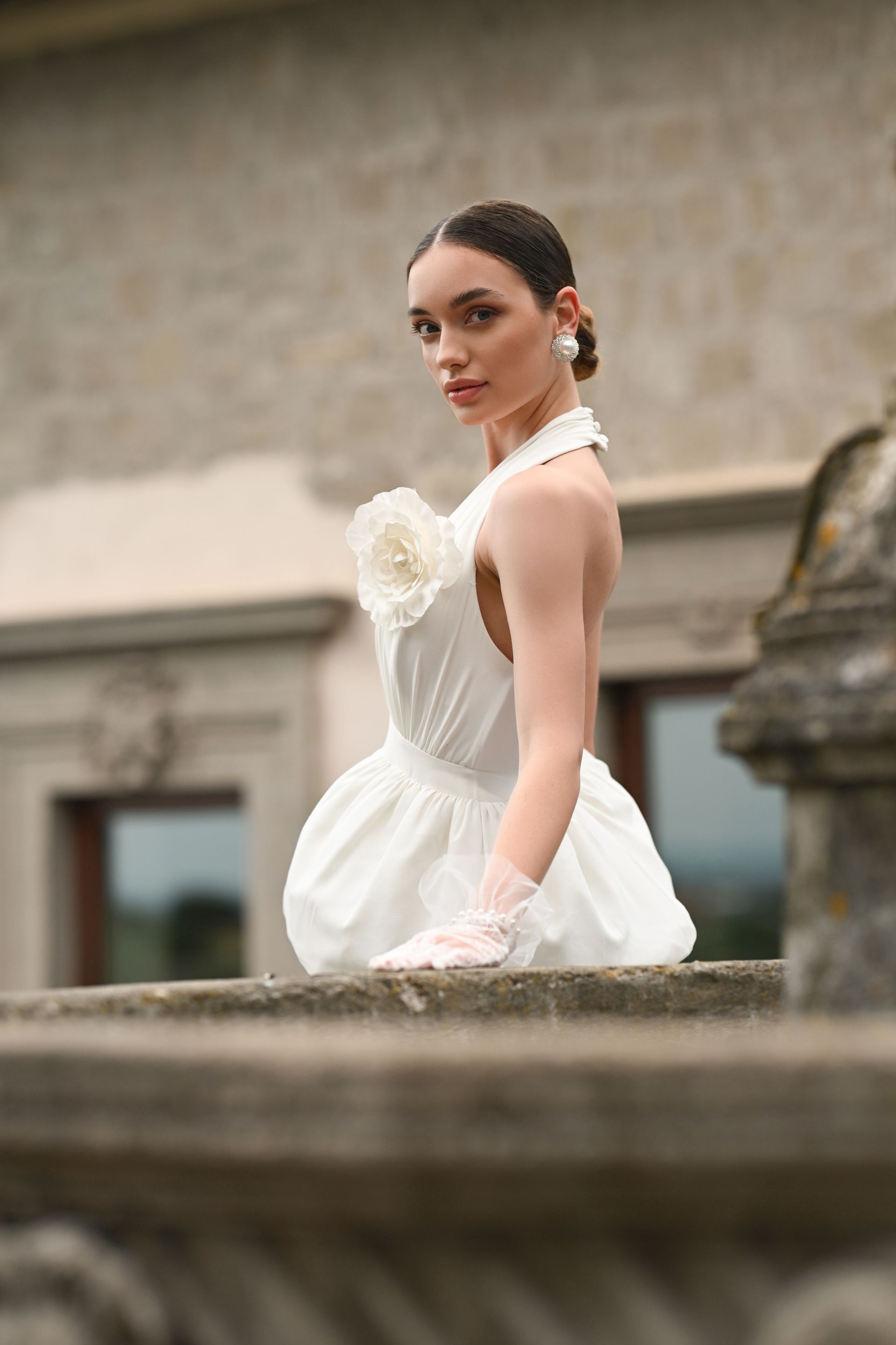 Woman in a white dress and gloves posing by a stone wall in an outdoor setting