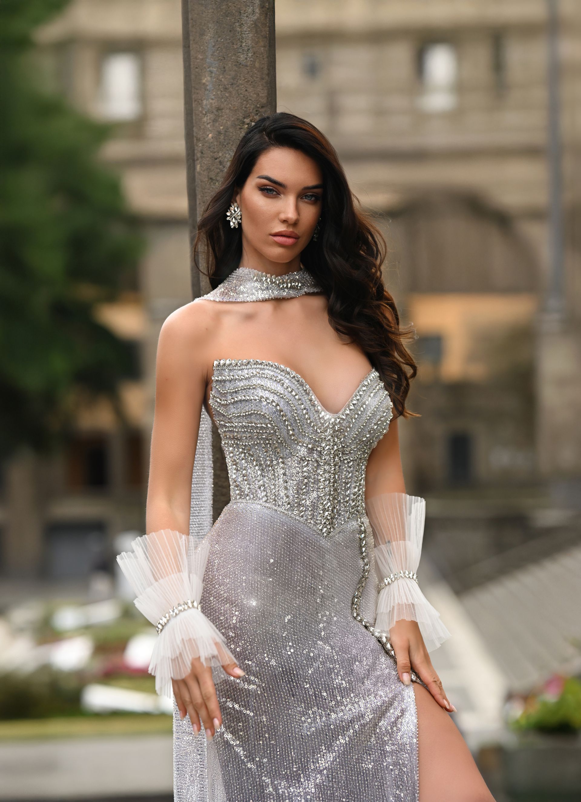 Woman in a silver evening gown with white gloves, standing outdoors in front of a blurred building.