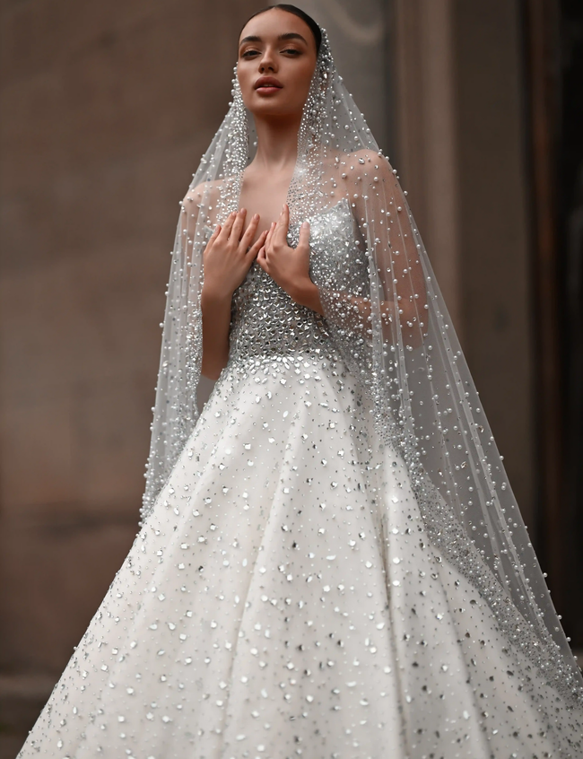 Bridal model in a white gown and sheer beaded veil standing in a softly lit stone setting