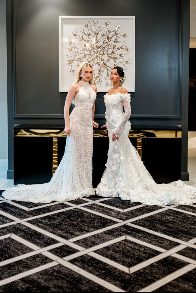 Two women in white wedding gowns pose indoors before a large floral artwork.