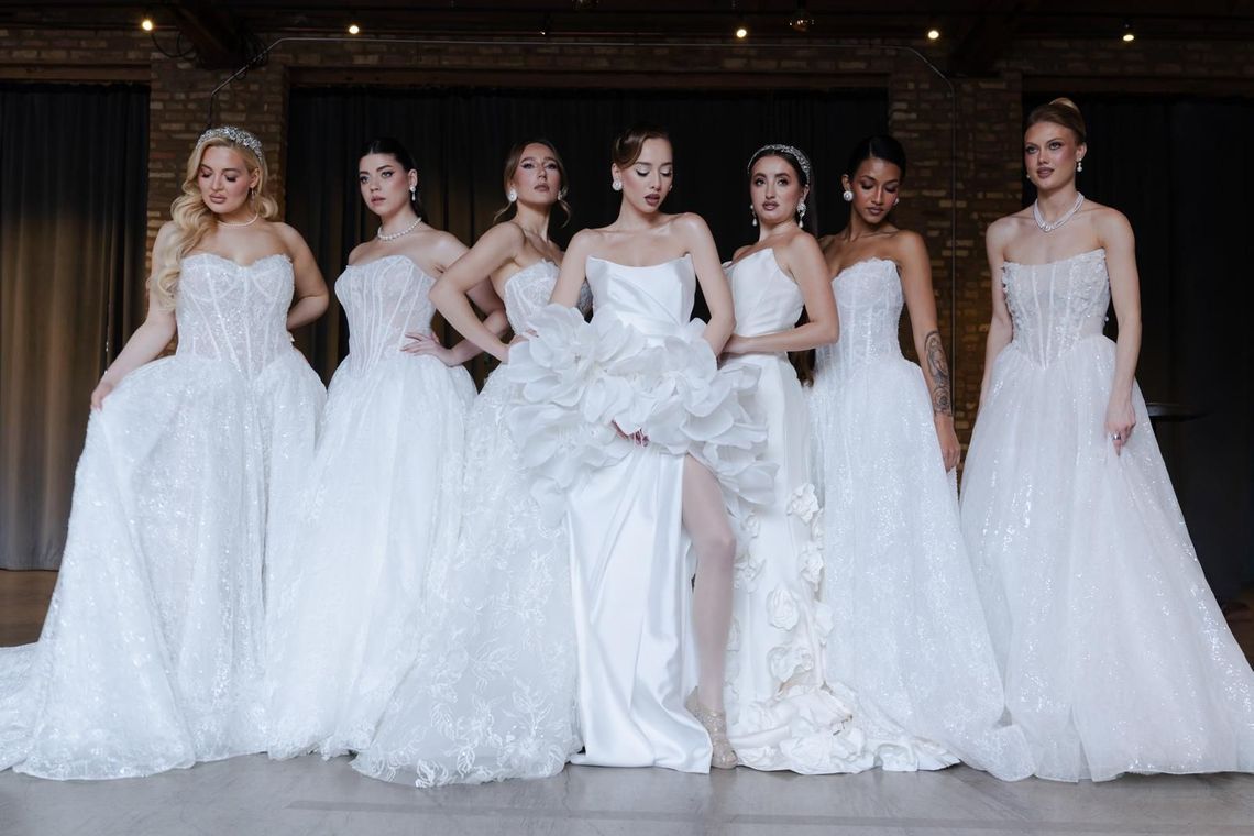 Seven women in white formal gowns posing together on a stage